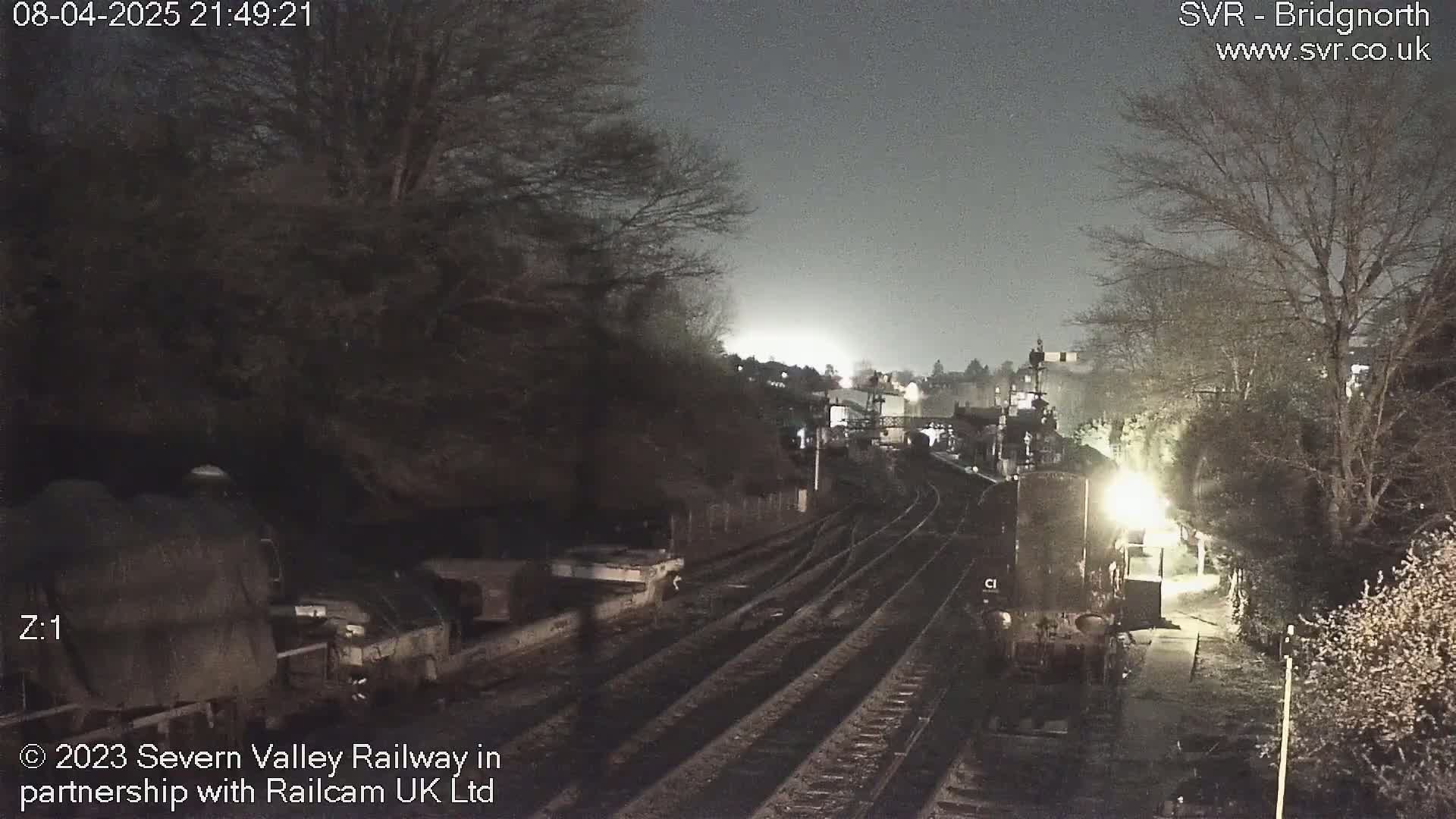 A nighttime view of a train station with train cars and tracks, under a dark, cloudy sky.