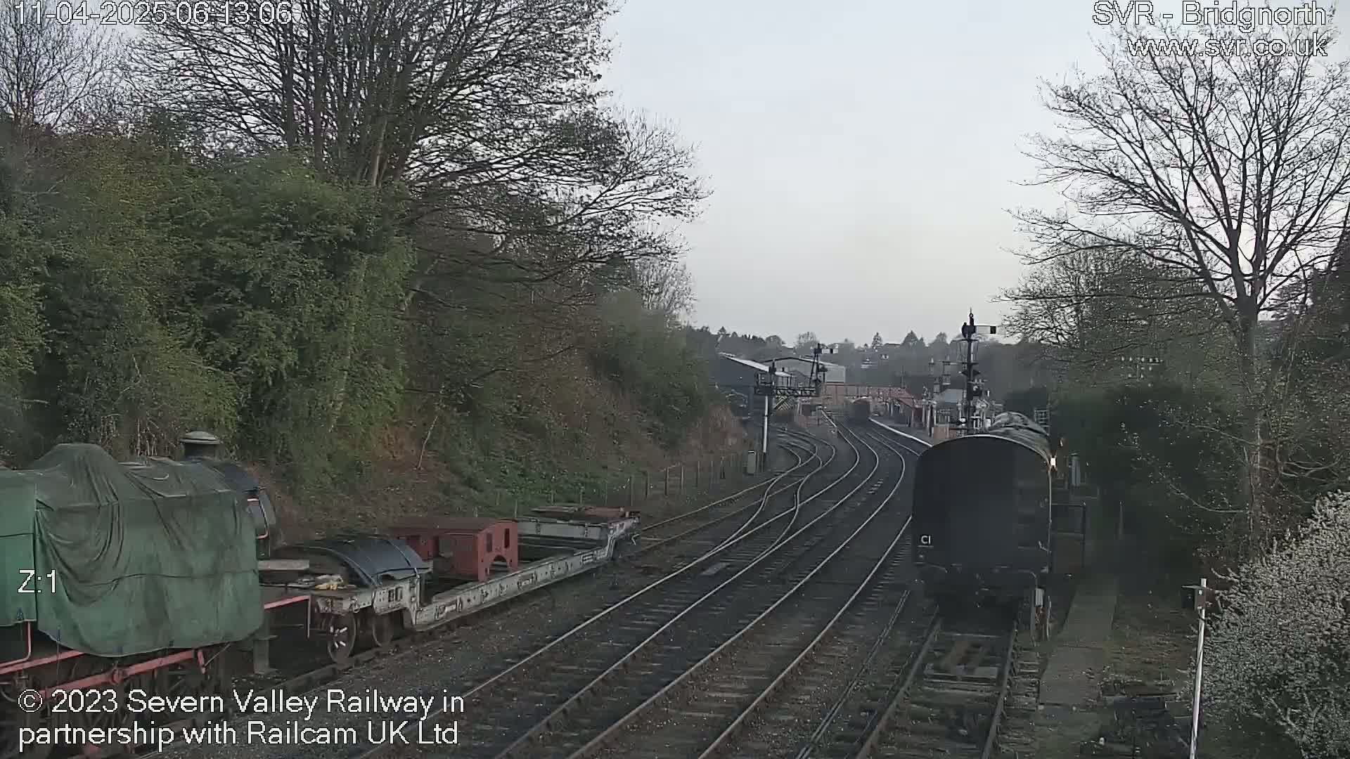 A dark-colored train is traveling on a railway track, passing a stationary train partially covered by a tarp, on an overcast day.