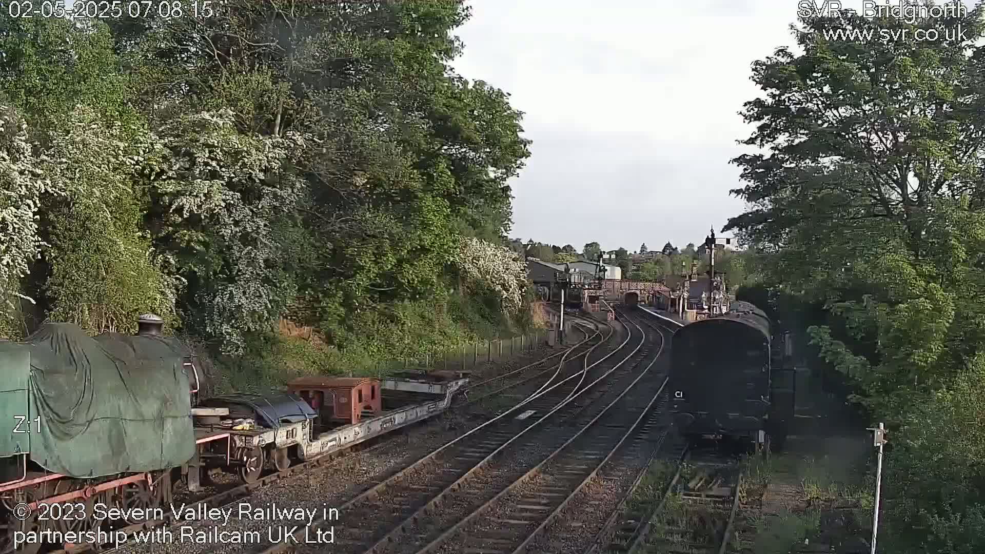 Two locomotives are visible on a railway track that curves through a lush green landscape under a partly cloudy sky.
