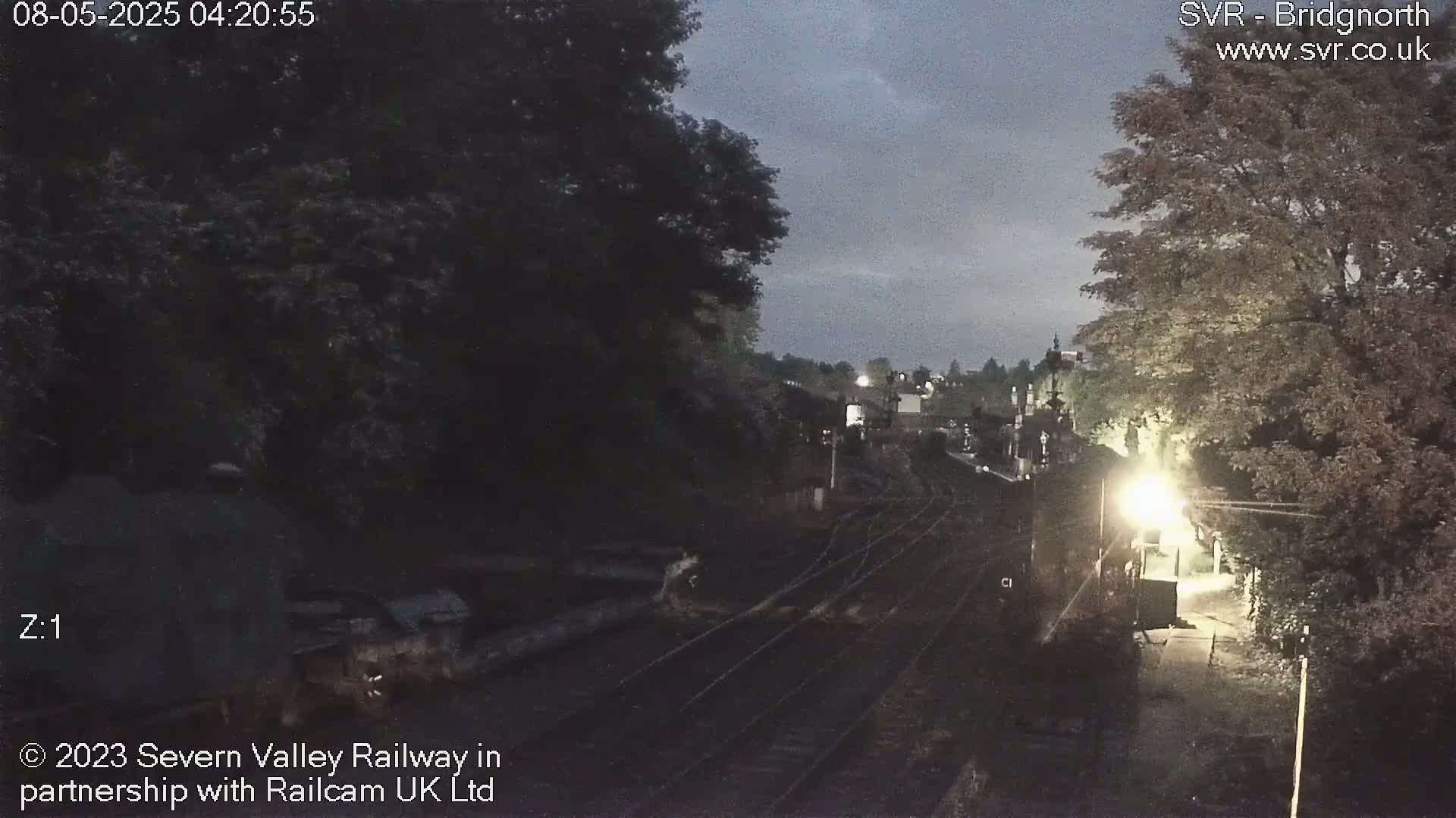 A dimly lit nighttime view of railroad tracks extending into a landscape of trees and buildings under a twilight sky.