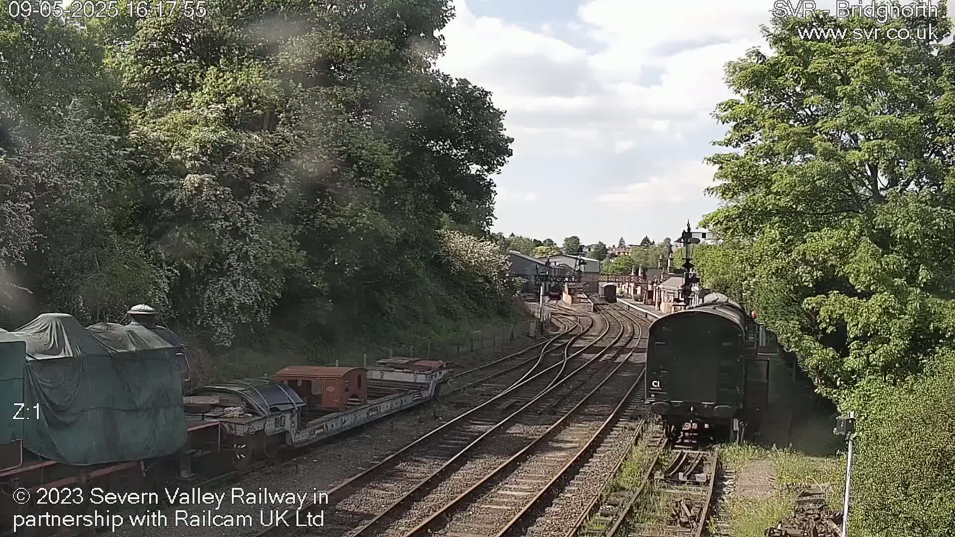 A partly sunny day shows a train on railway tracks curving through a lush green landscape, with other railcars and a covered locomotive visible nearby.