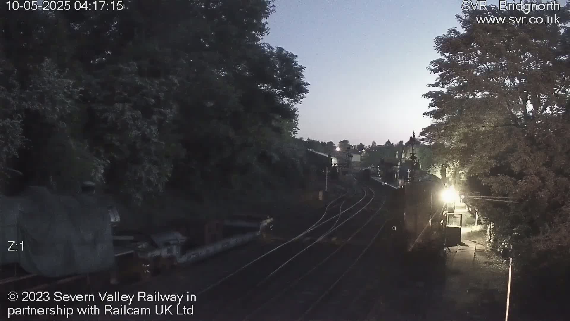 A twilight view of railroad tracks curving through a landscape of trees and railway structures.
