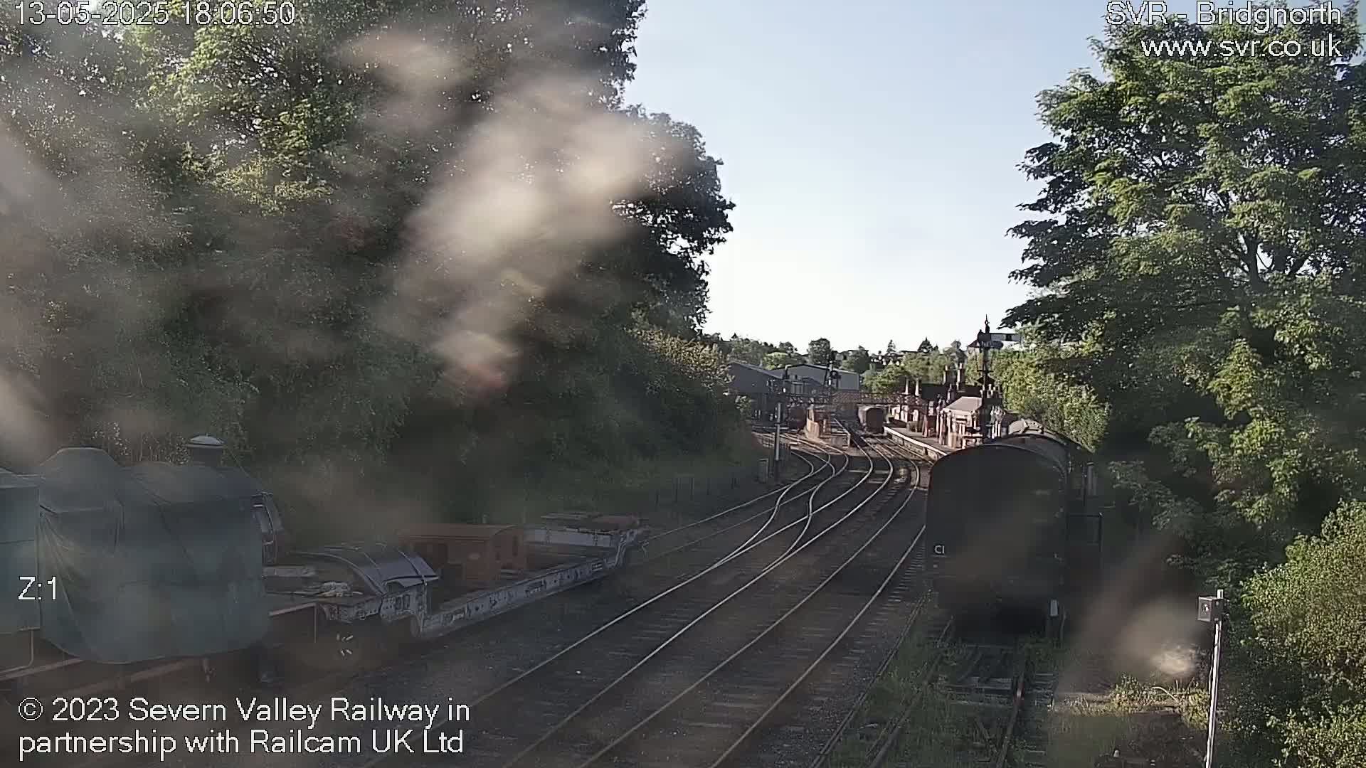 A steam train is partially visible near a railway station on a sunny day, with smoke emanating from the train and surrounding trees.