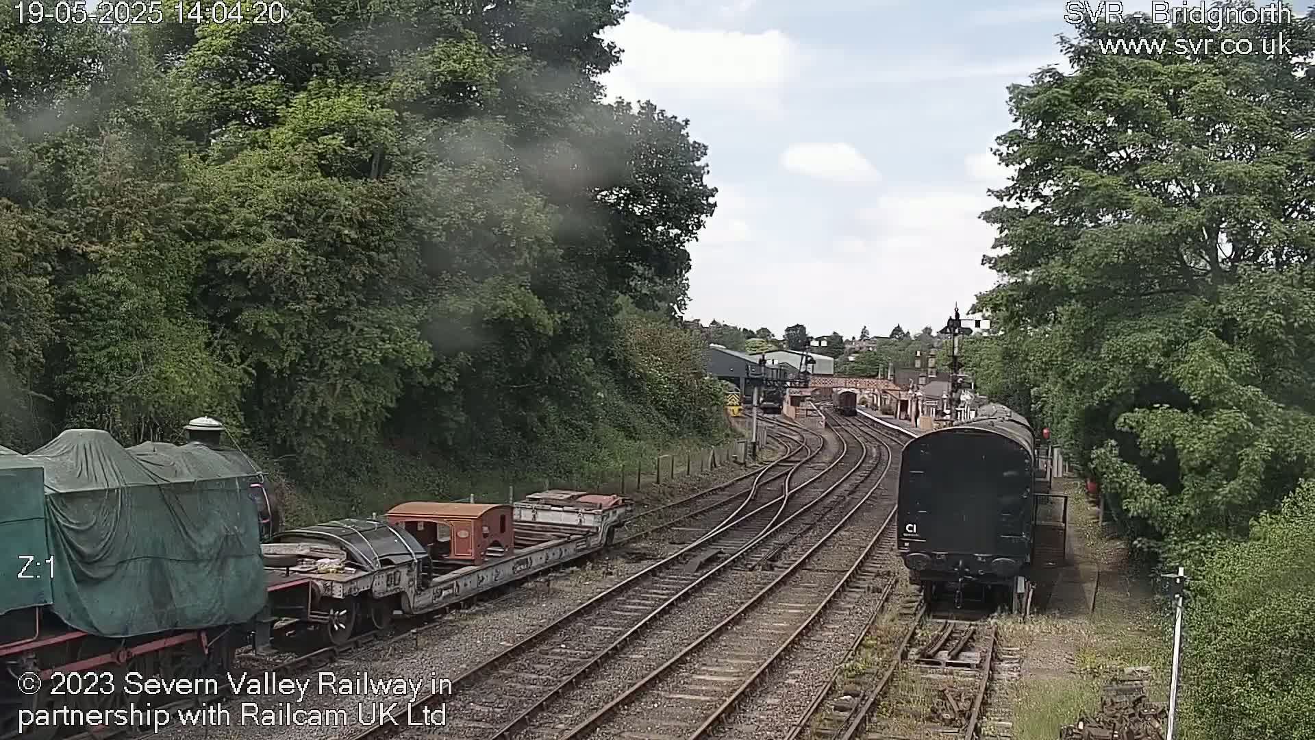 A partly cloudy day shows a train yard with multiple train cars, some covered, on railway tracks surrounded by lush green trees.