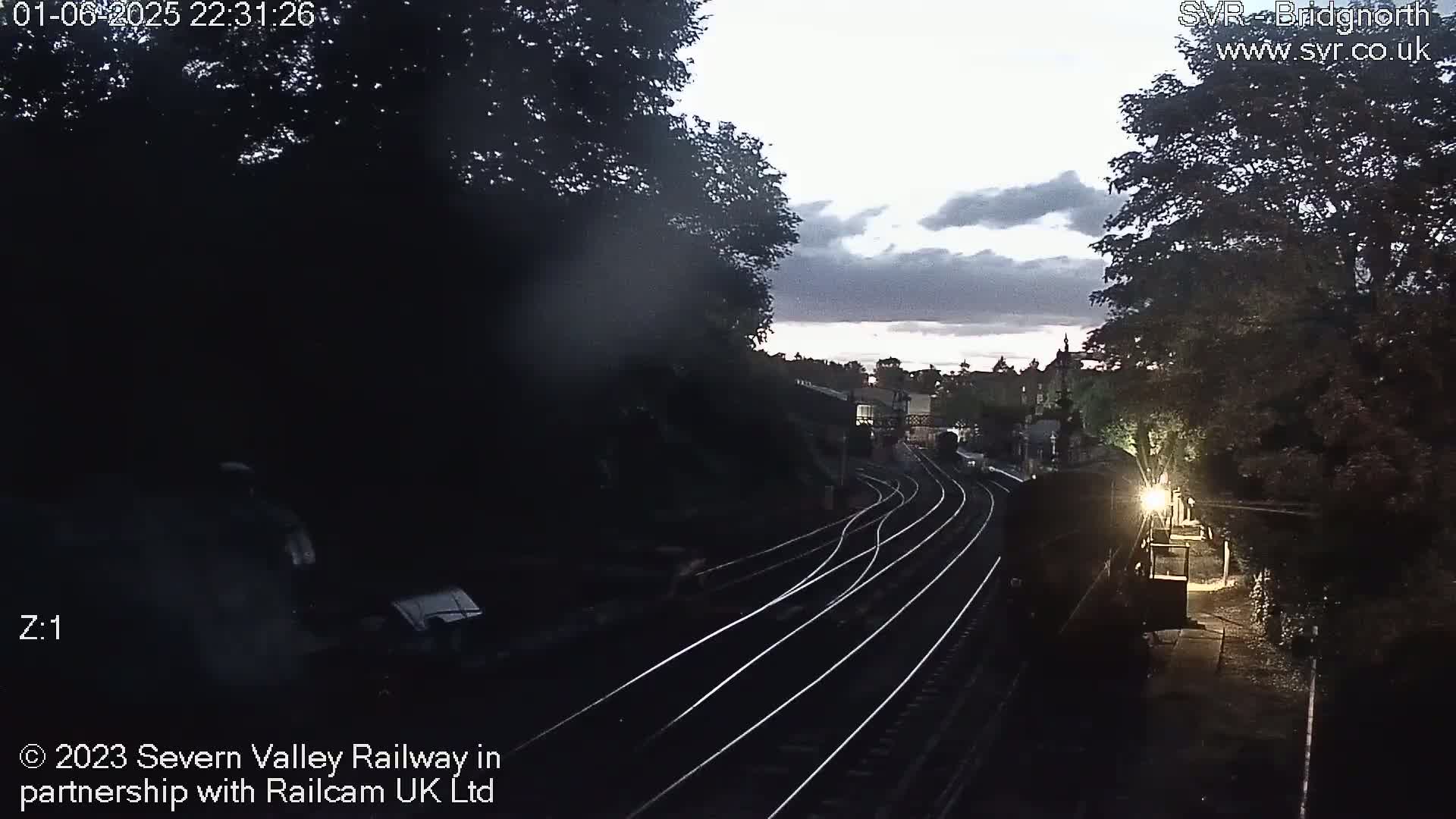A train approaches a dimly lit station at twilight under a partly cloudy sky.
