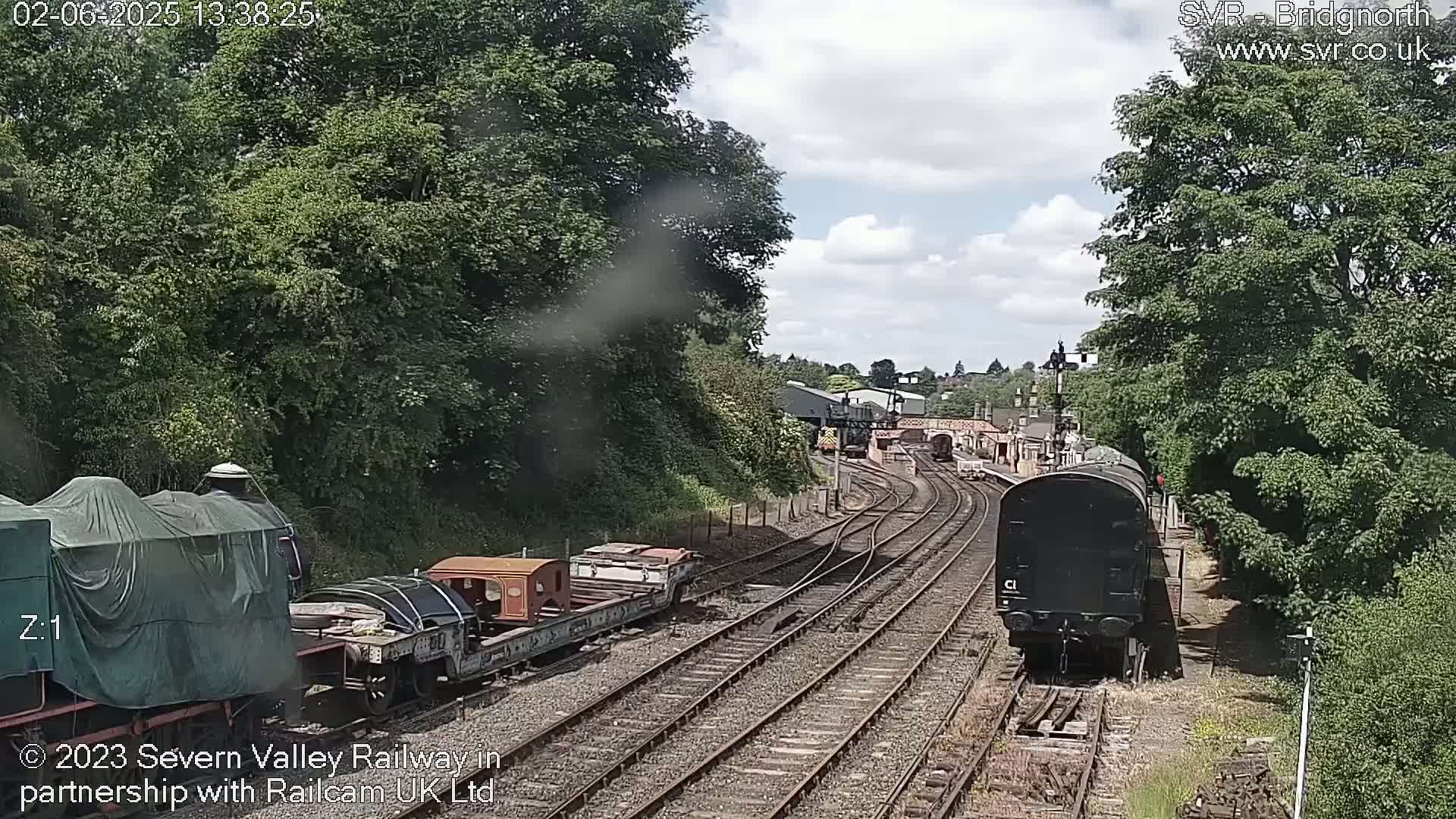 A partly cloudy day at a train yard shows multiple train cars on several tracks, with lush green trees surrounding the area.