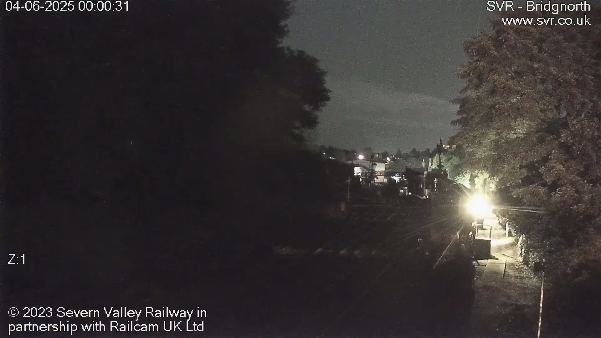 A nighttime view of a dimly lit path leading toward buildings and trees under a dark sky.