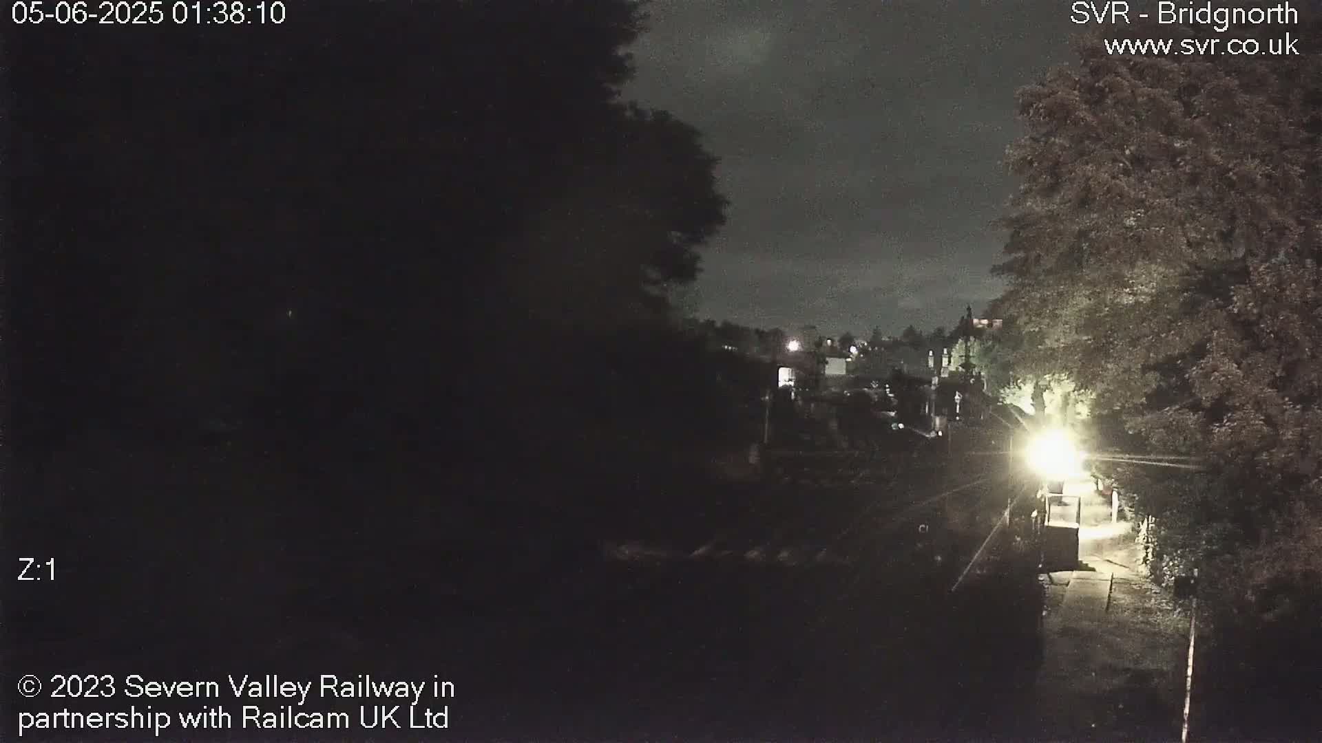 A nighttime, dimly lit view of a path leading toward a brightly lit area near some trees and buildings.