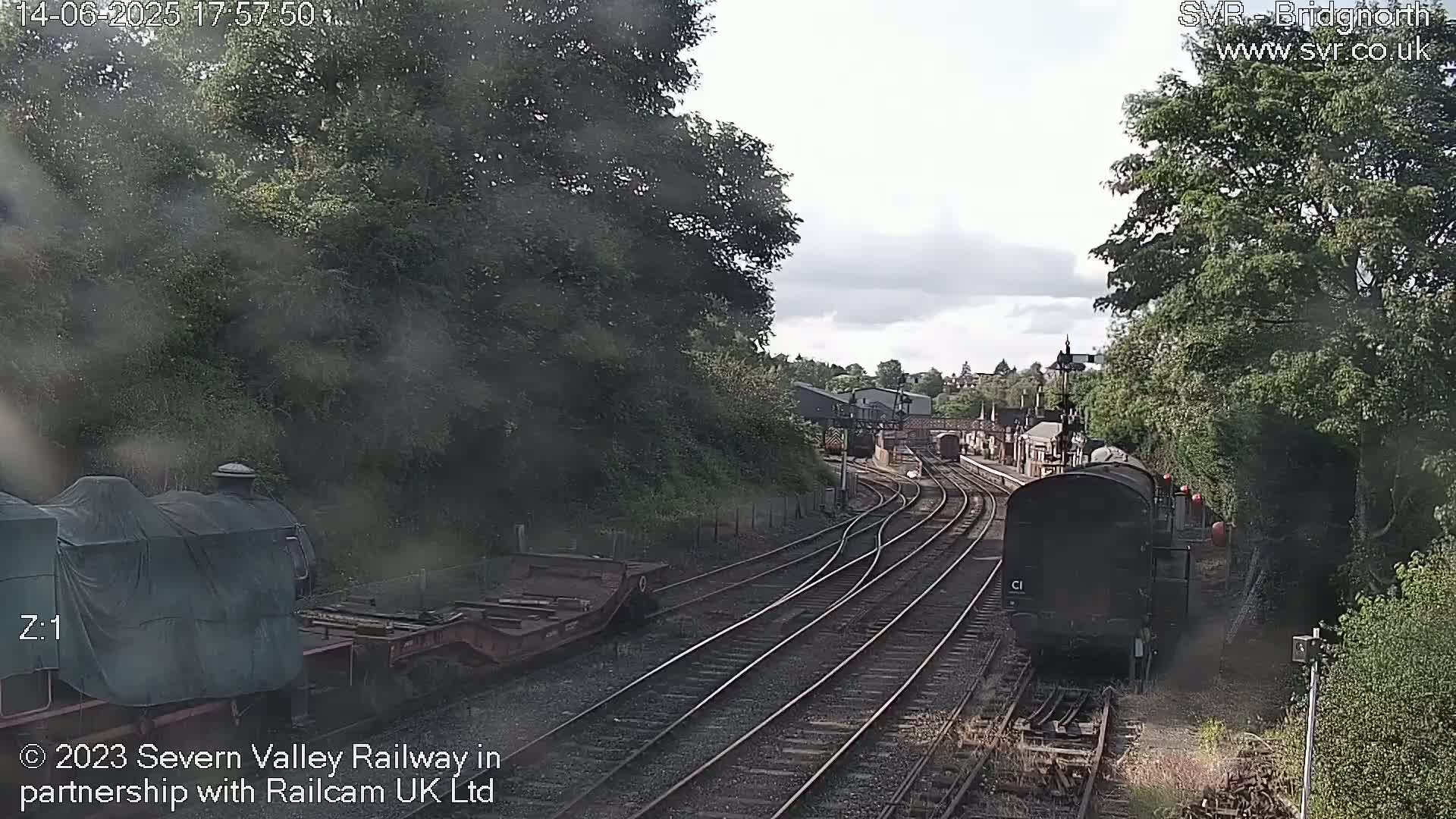 A view from a train shows multiple train tracks curving through a wooded area under a partly cloudy sky, with a dark-colored train car visible in the foreground.