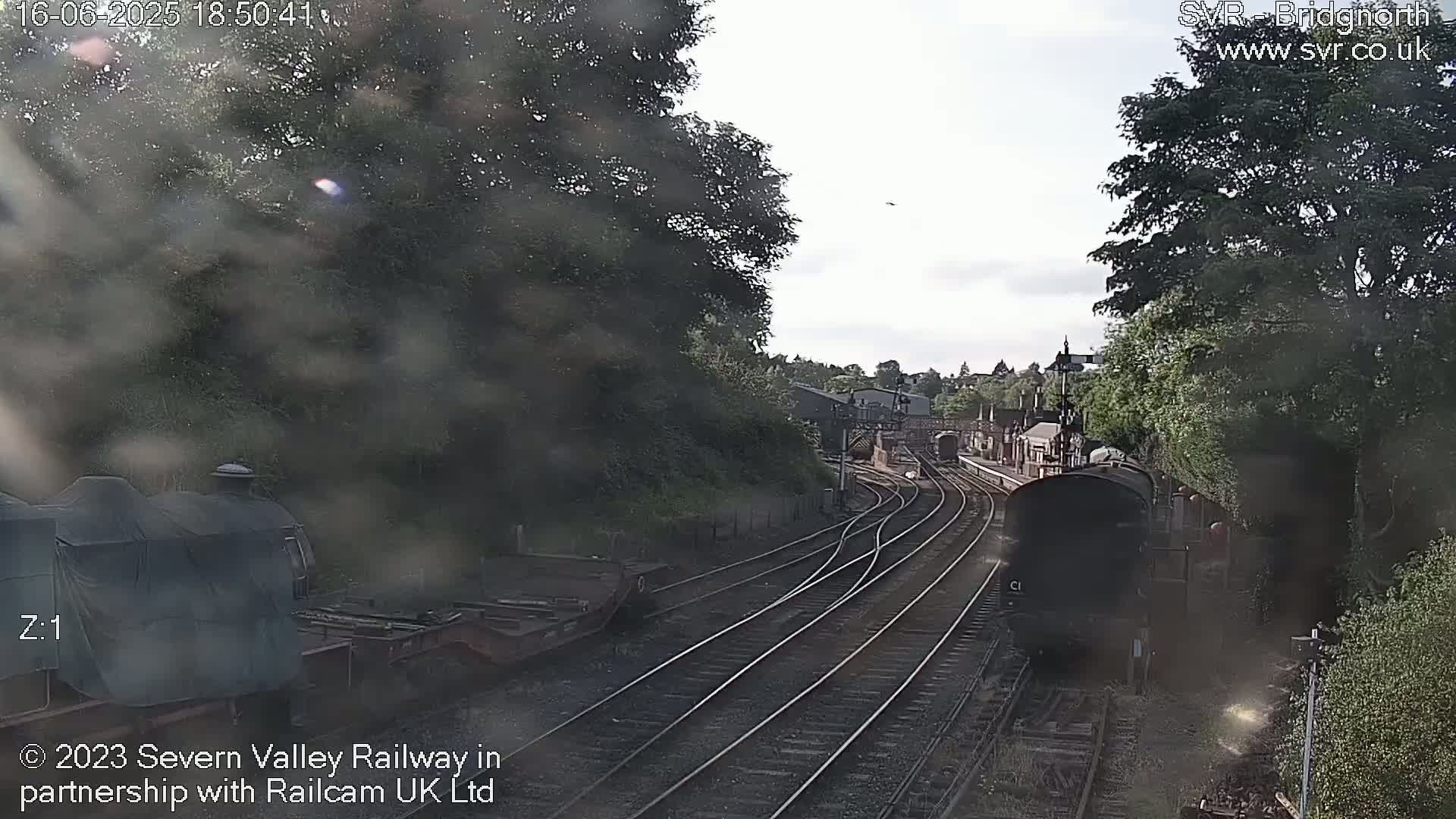 A view from a train shows railway tracks branching off, a dark-colored train car moving along one track, and a partly visible train covered in a tarp, all under a mostly clear sky.