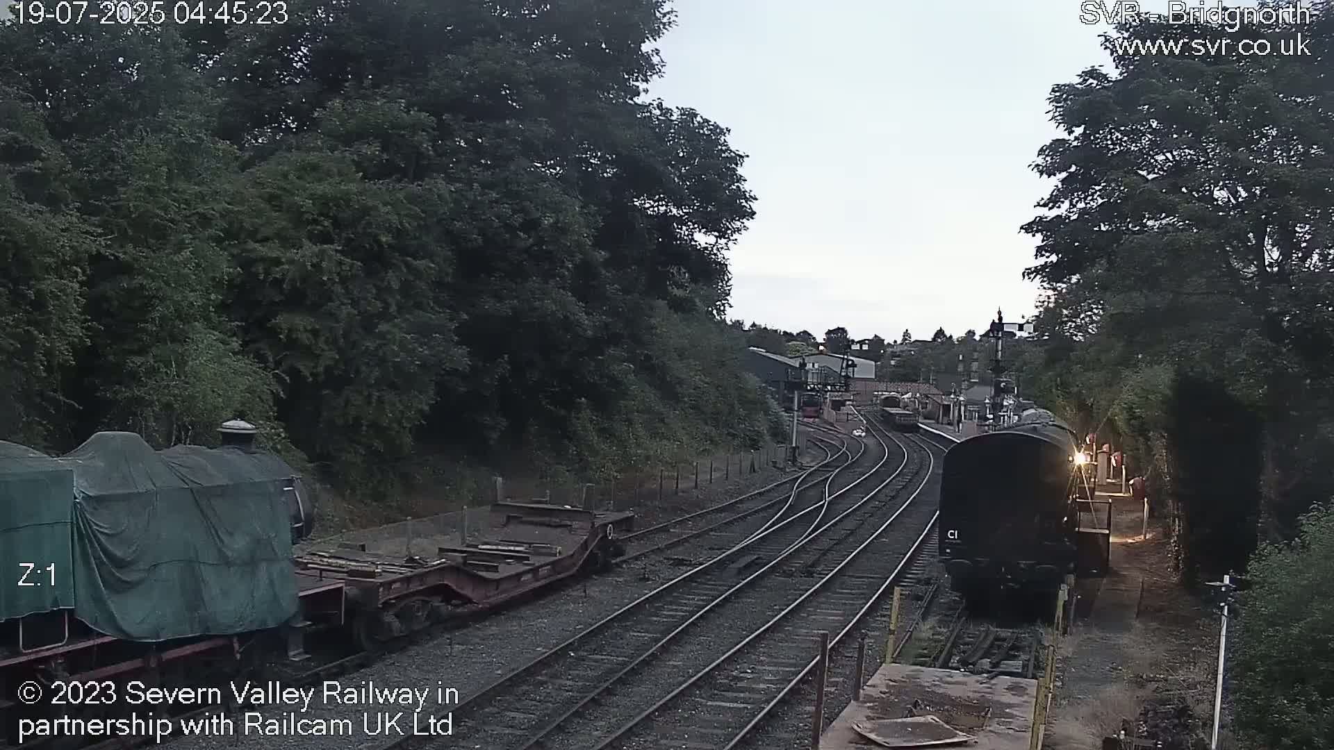 A train approaches a railway station on a slightly overcast day, passing other railway cars and tracks.