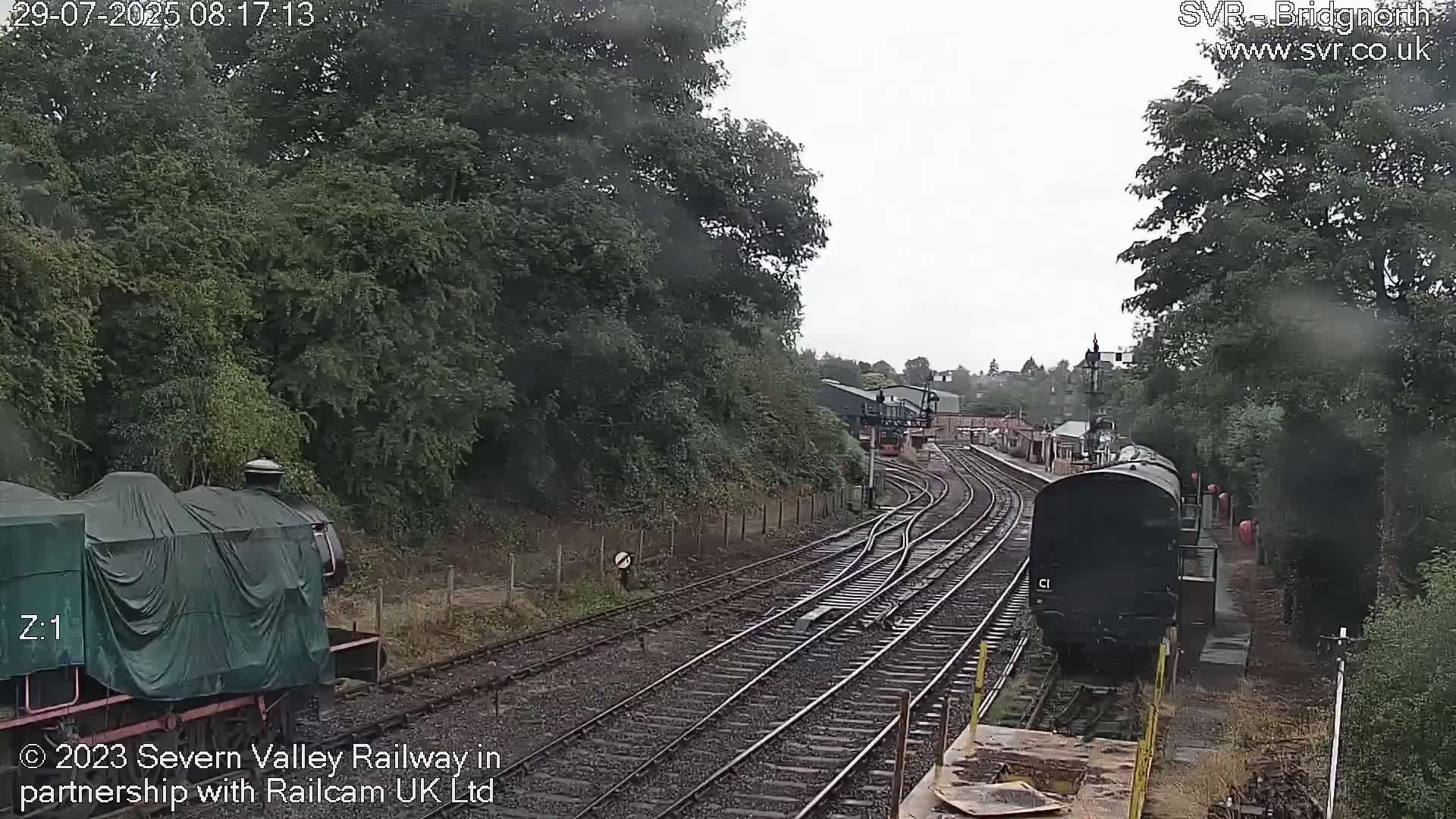 A view of a railway line on an overcast day, with two trains visible; one covered and stationary, the other dark-colored and moving.