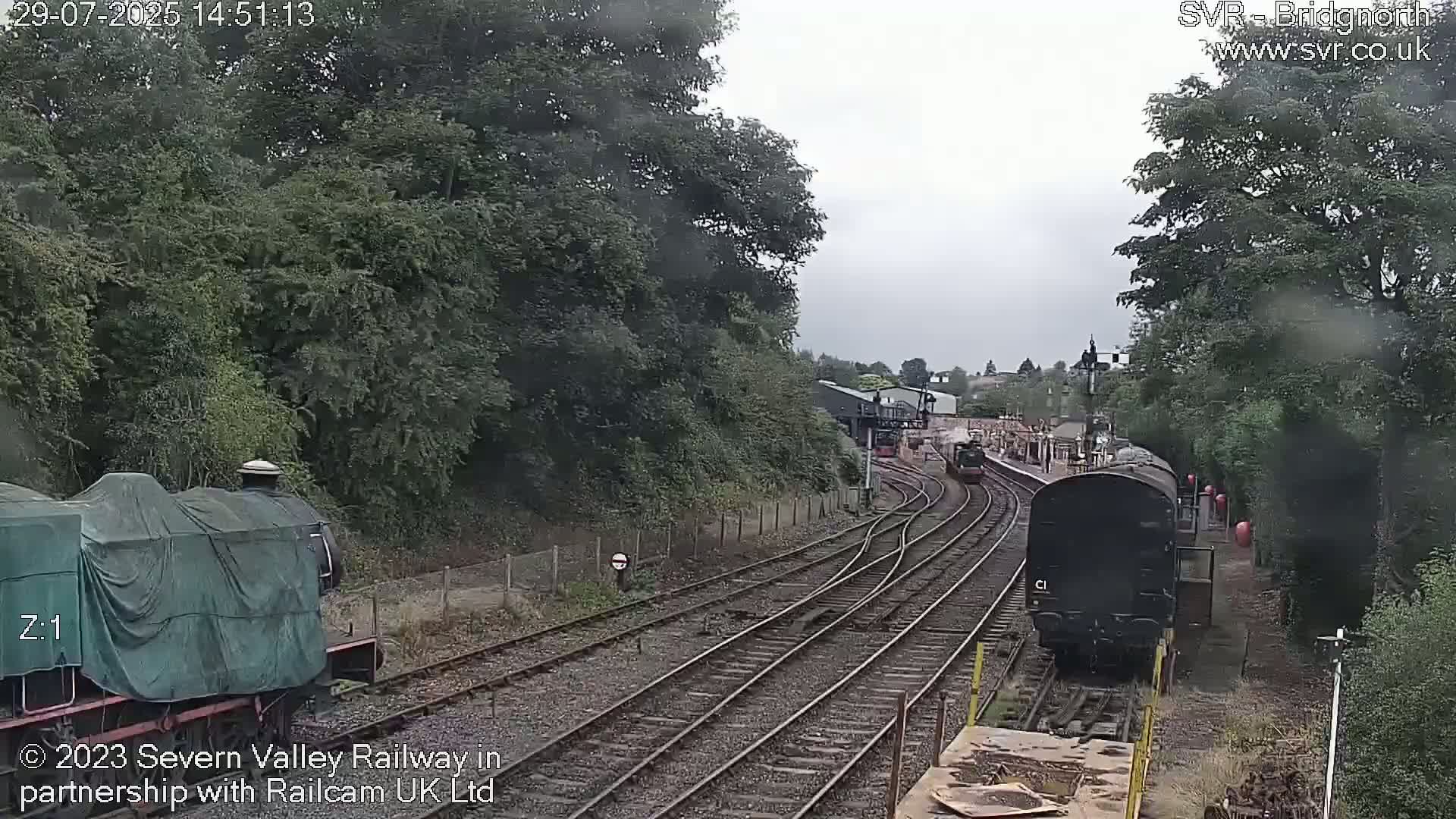 A view of a railway line on an overcast day, with two trains visible; one covered and stationary, the other dark-colored and moving.