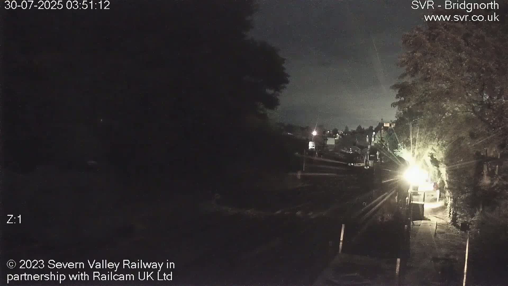 A view of a railway line on an overcast day, with two trains visible; one covered and stationary, the other dark-colored and moving.