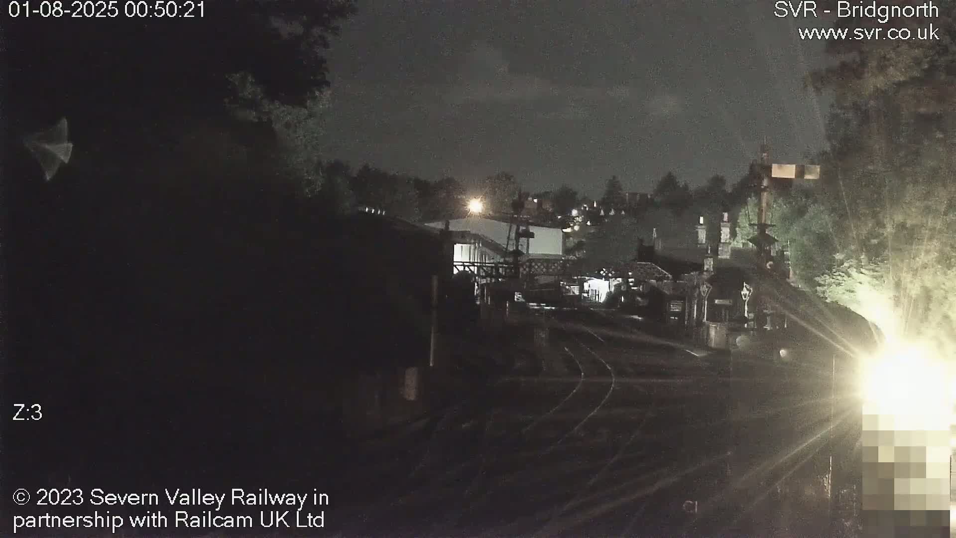 A nighttime view of a train station with train tracks and buildings under a dark, cloudy sky.