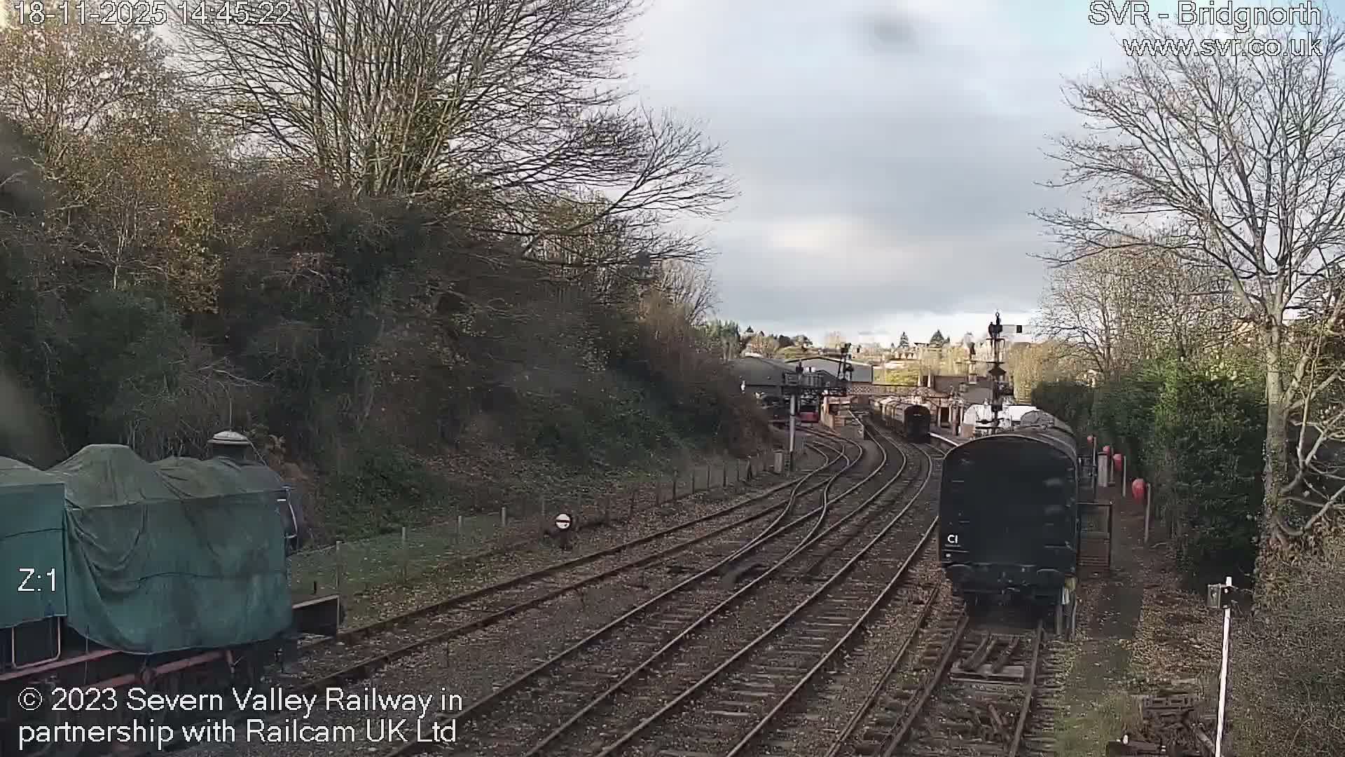 A nighttime view of a train station with train tracks and buildings under a dark, cloudy sky.