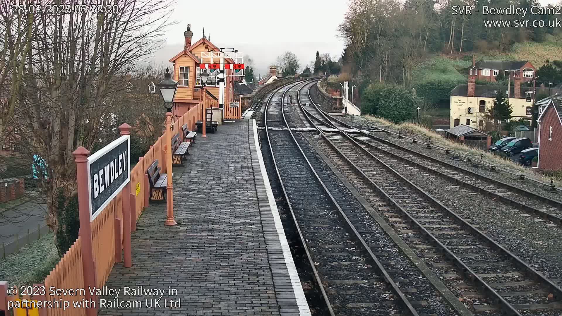 A train station platform with multiple tracks curves gently into the distance under an overcast sky.