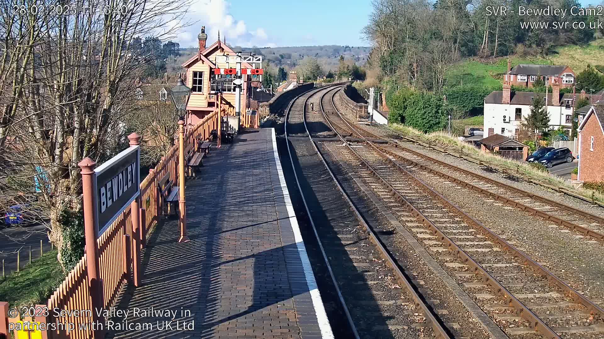 A sunny view of a train station platform with multiple train tracks extending into the distance, past a signal box and residential buildings.