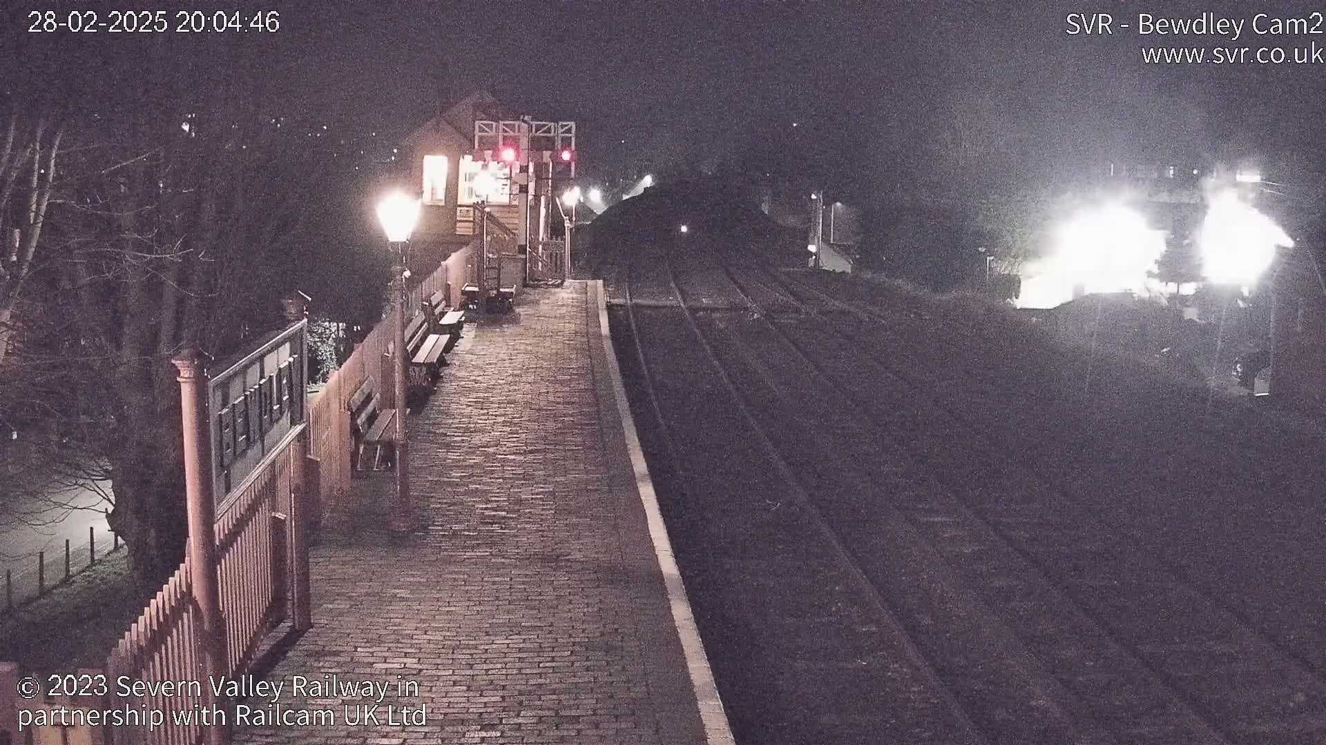 A nighttime view of a train station platform with a train approaching, illuminated by lampposts and the train's headlights.