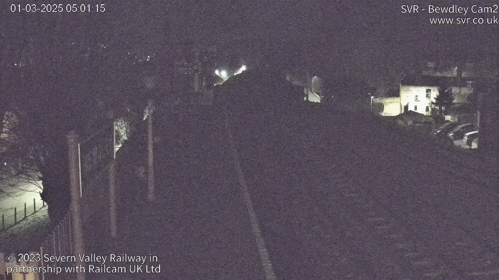 A nighttime, low-light view of a railway track and buildings, possibly a station, with some fog or mist present.
