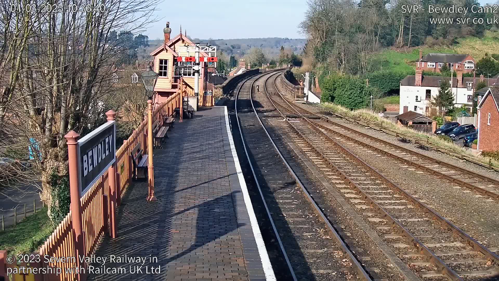 A sunny view of a train station platform with multiple train tracks extending into the distance, past houses and trees.