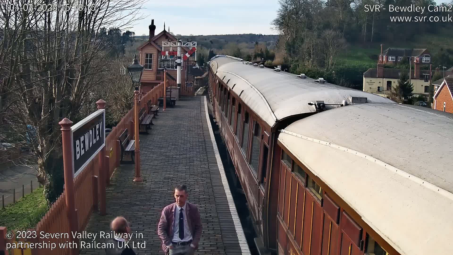 A long, dark-red train is at a station on a sunny day, with a man in a suit standing on the platform.