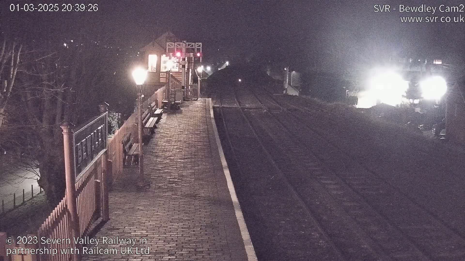 A brick pathway at a train station at night is illuminated by a lamppost, with a train approaching in the distance under dark, nighttime conditions.