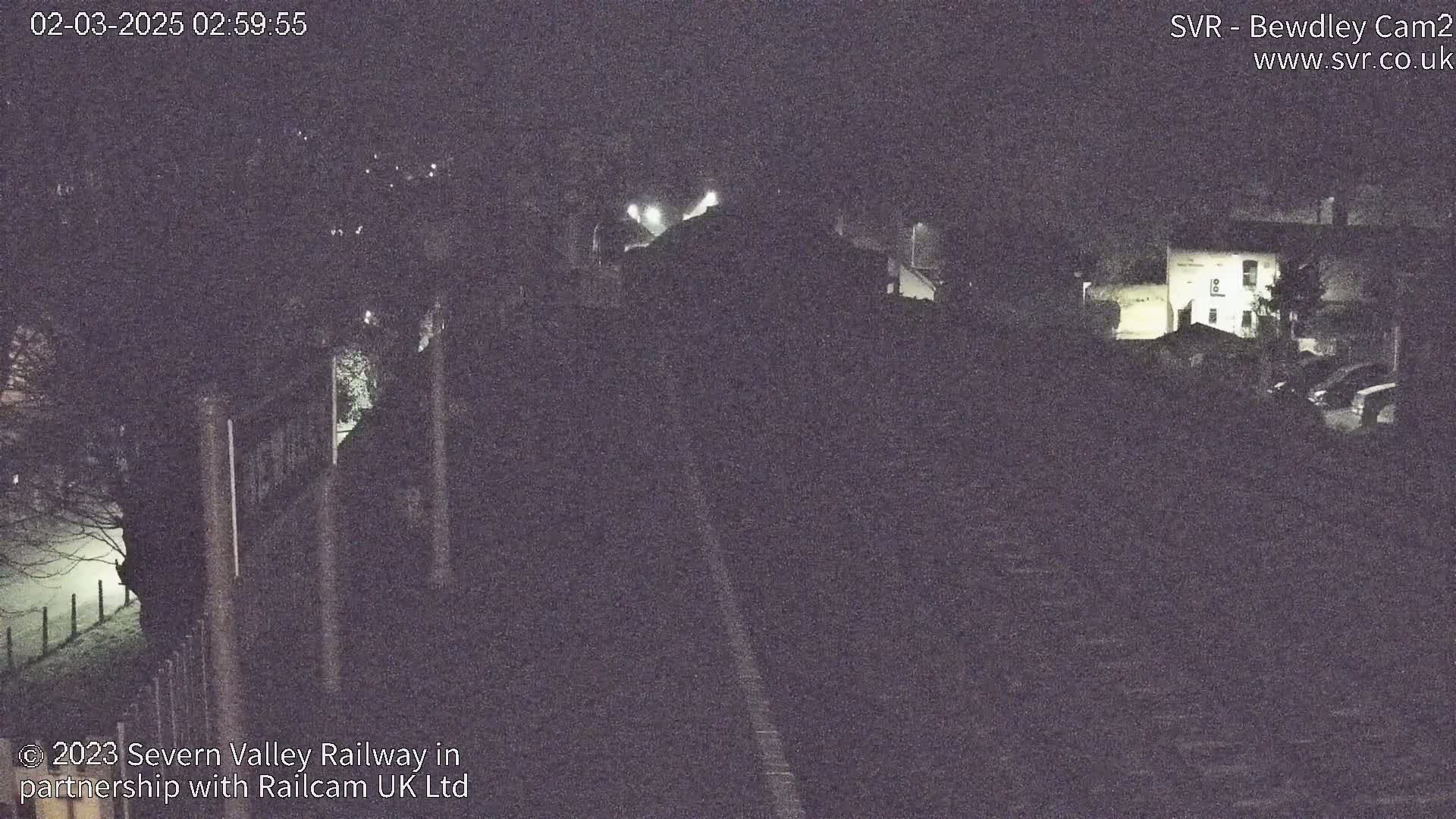 A nighttime, dimly lit view of a railway track and surrounding buildings, possibly a station, under slightly foggy conditions.