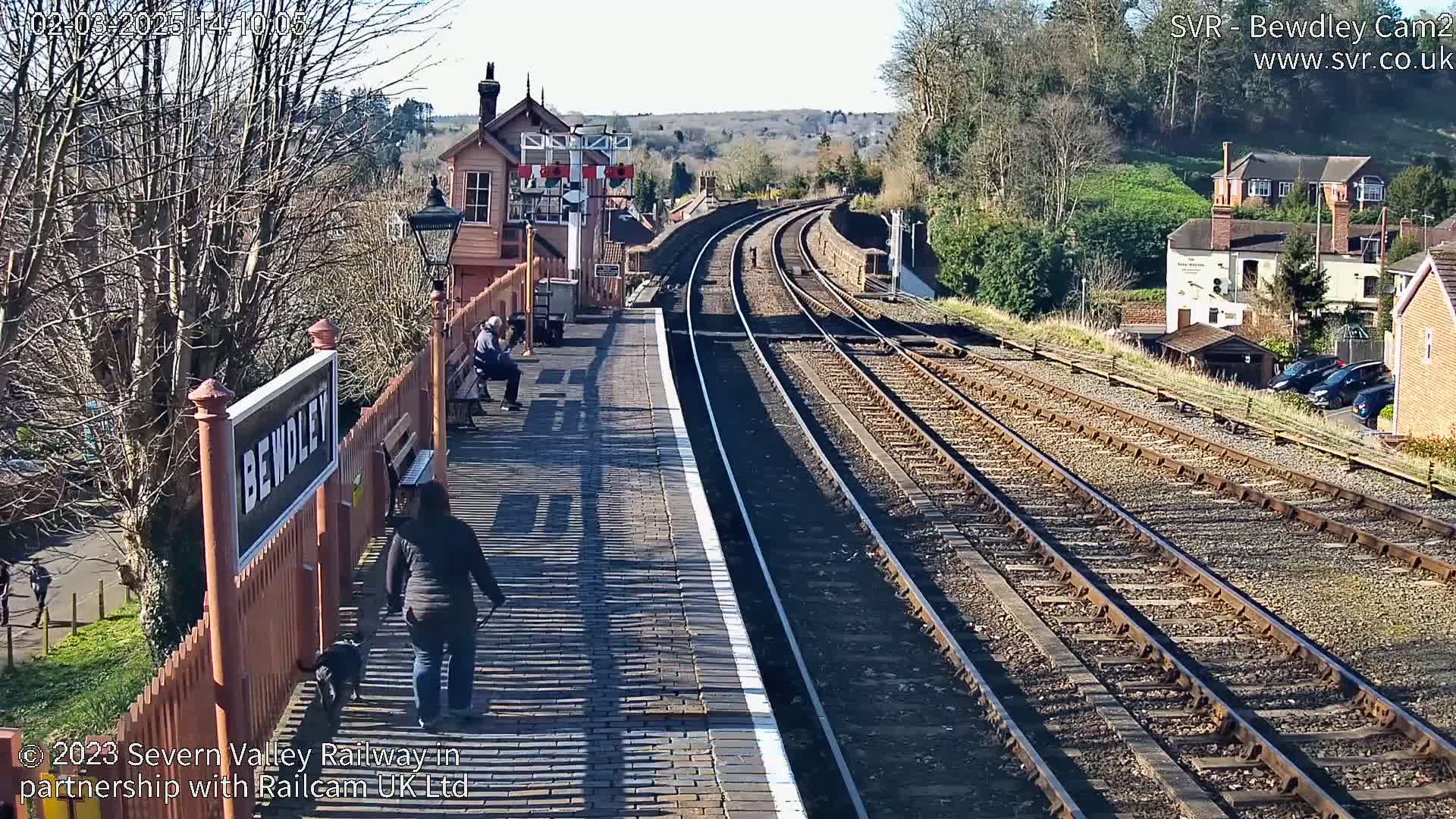 A sunny day at a train station shows a brick platform with multiple train tracks extending into the distance, alongside a small building and a person walking a dog.