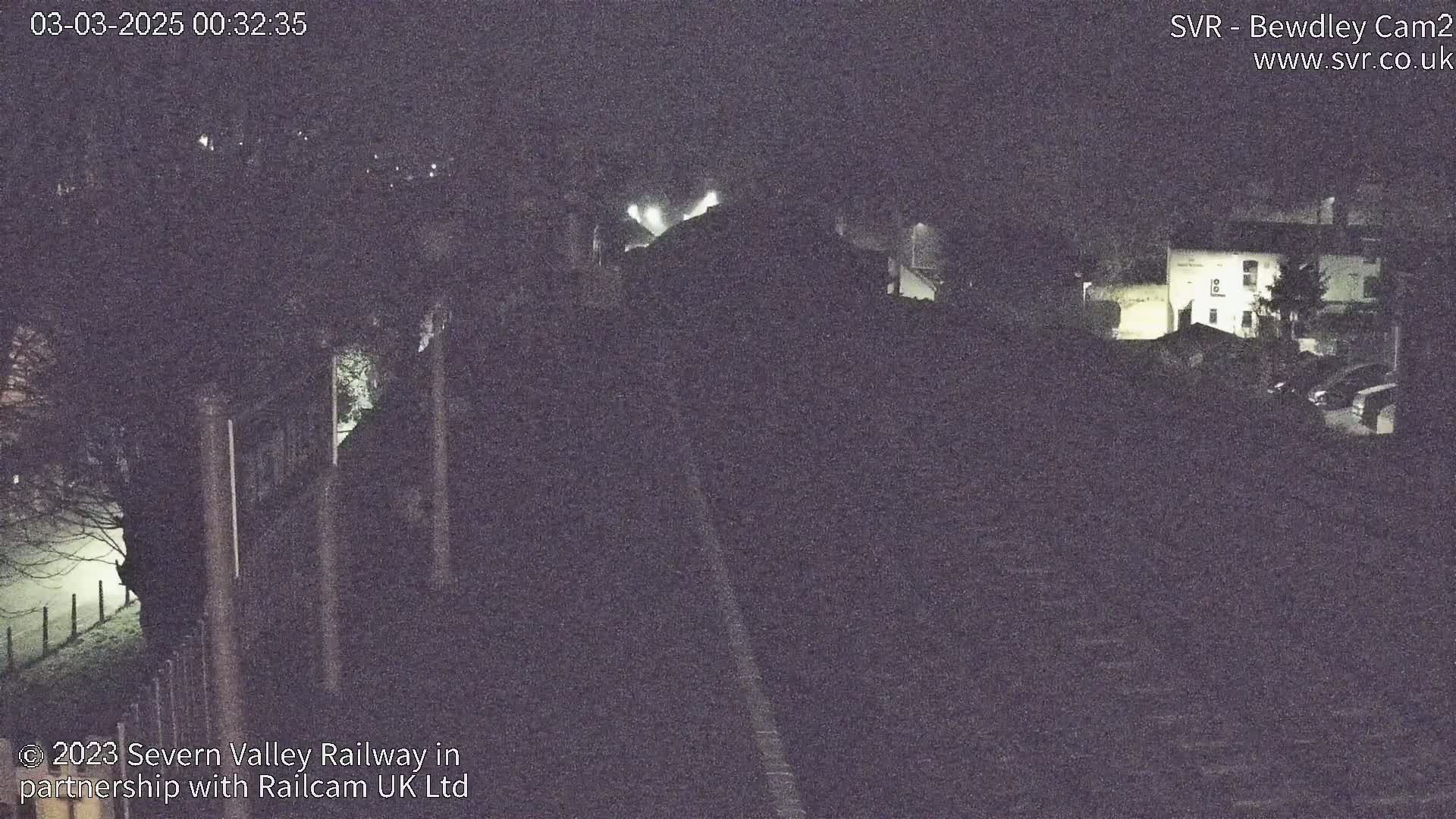A nighttime, low-light view of a railway track and buildings, possibly a station, with some fog or mist in the distance.