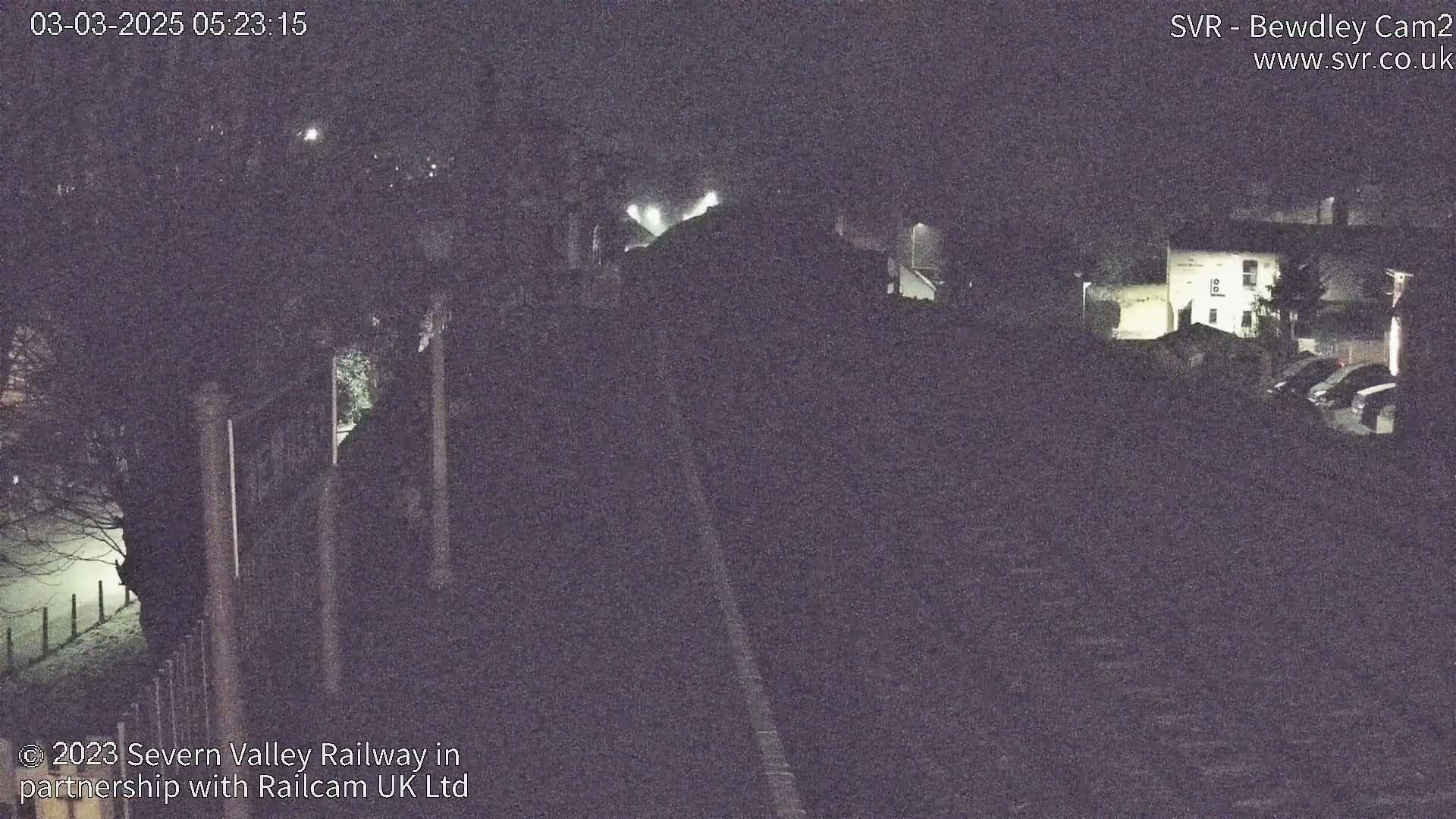A nighttime, low-light view of a railway line, a building, and some parked cars, possibly in foggy conditions.