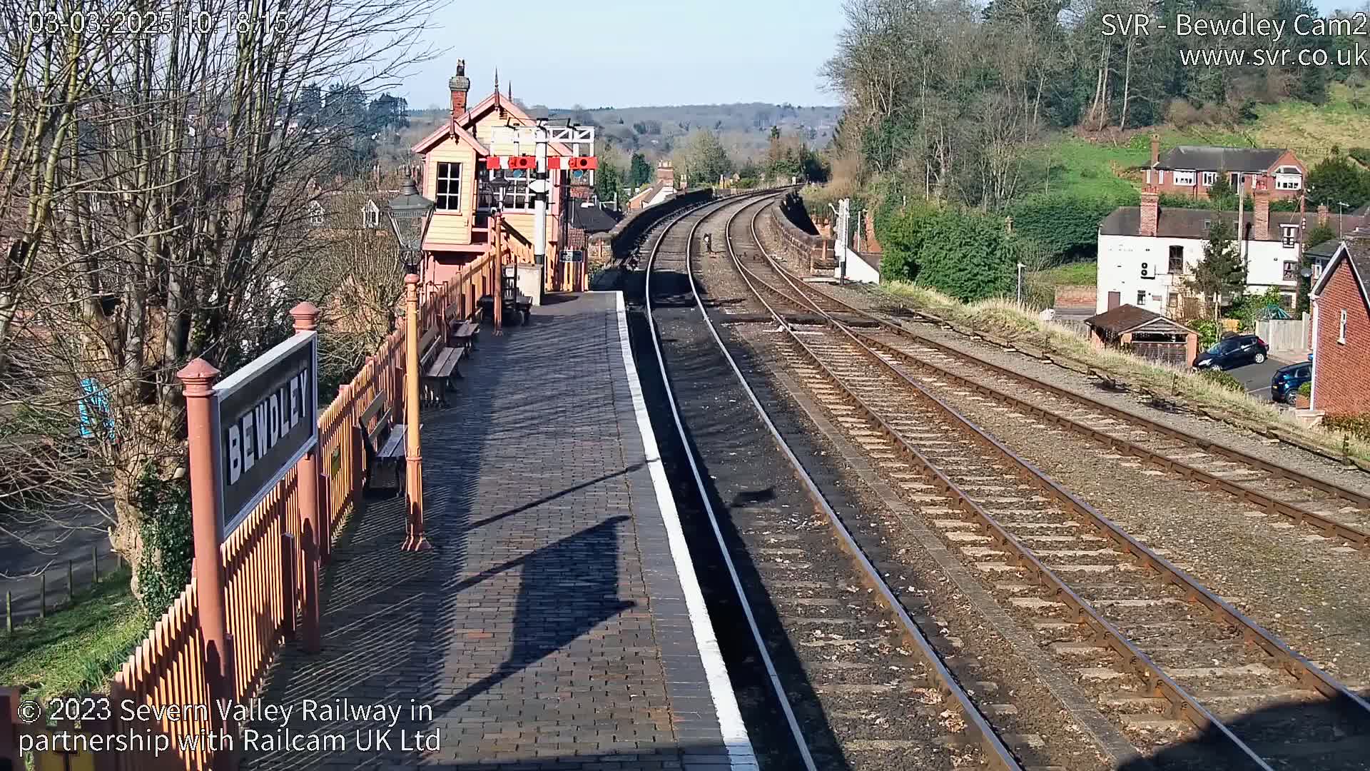 A sunny view of a train station platform with a signal box, railway tracks extending into the distance, and houses visible beyond the tracks.
