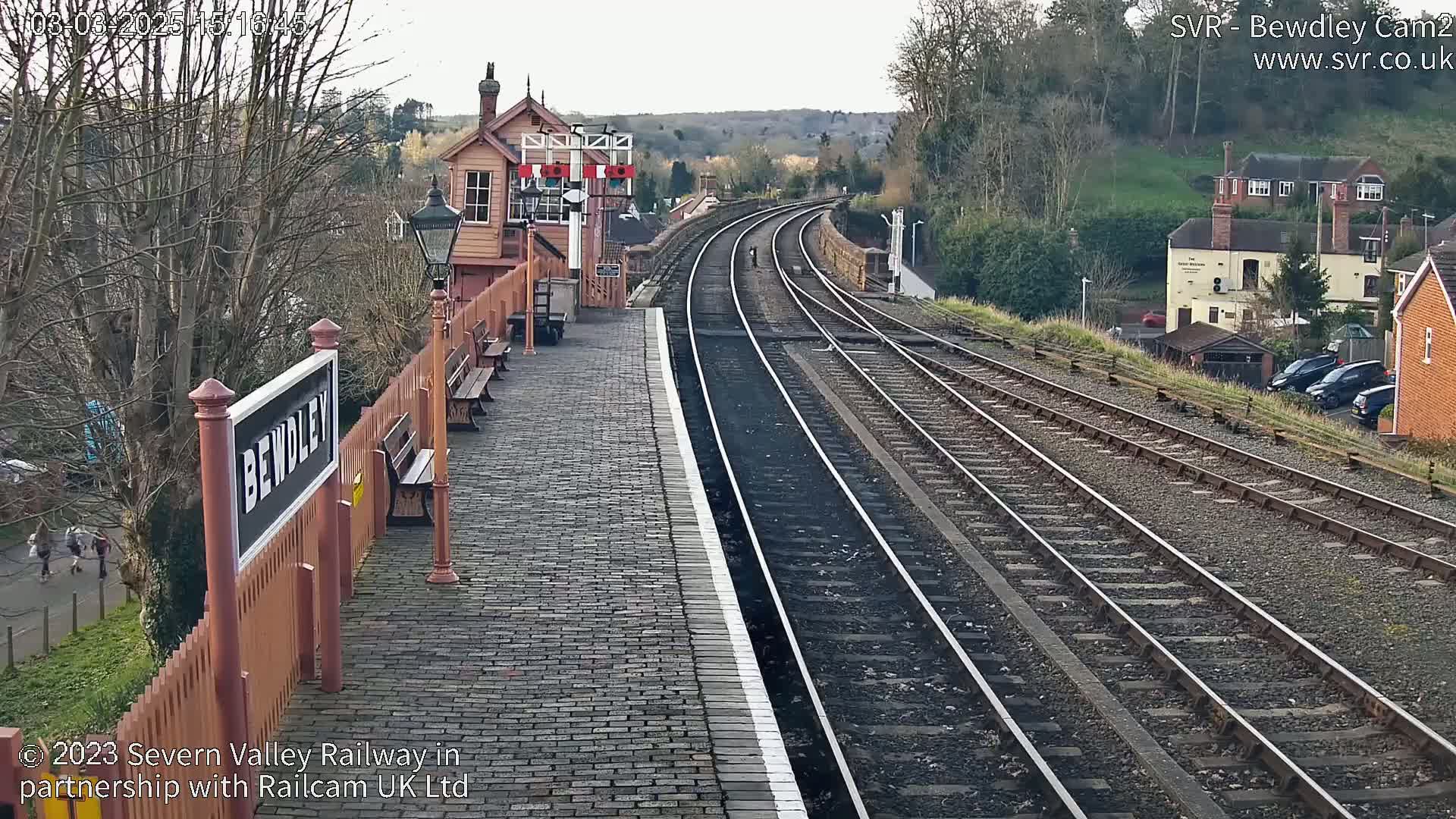 A train station platform with brick pavement and multiple train tracks extends into the distance under an overcast sky.