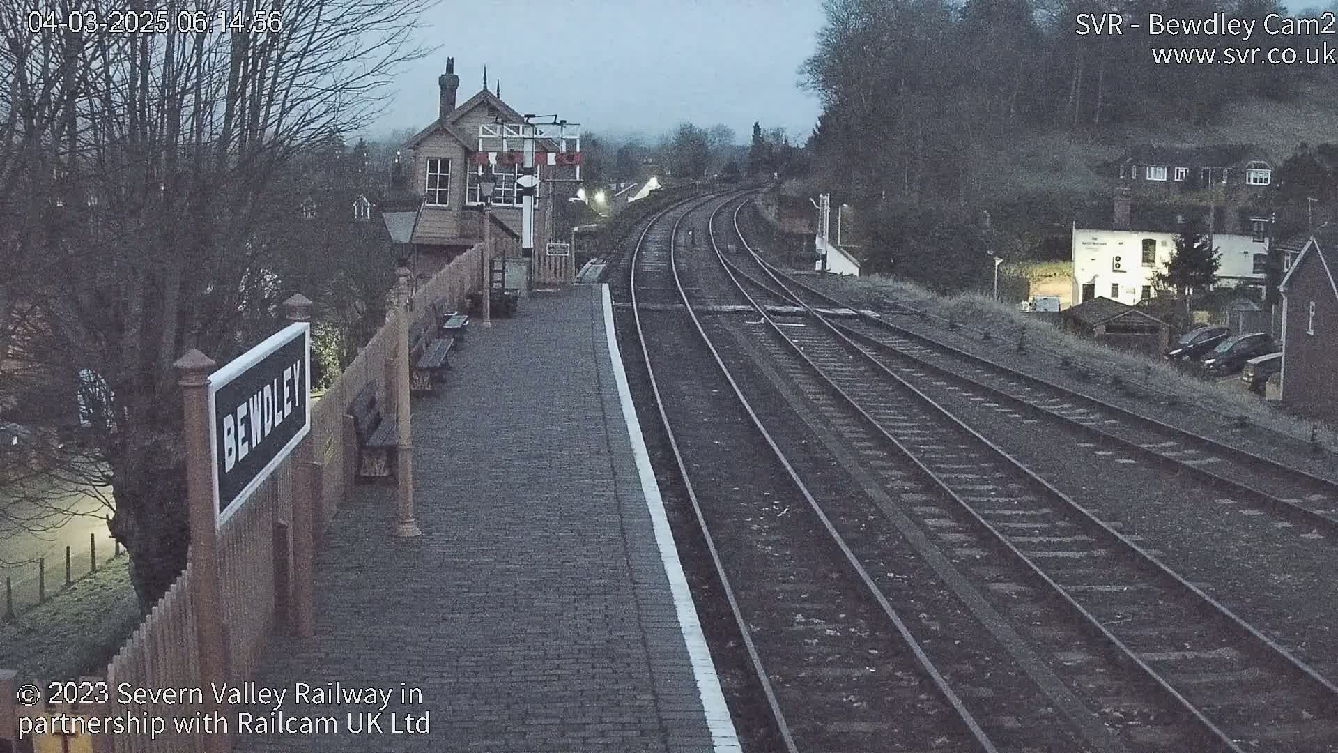 A train station platform with multiple train tracks extends into the distance under an overcast sky.
