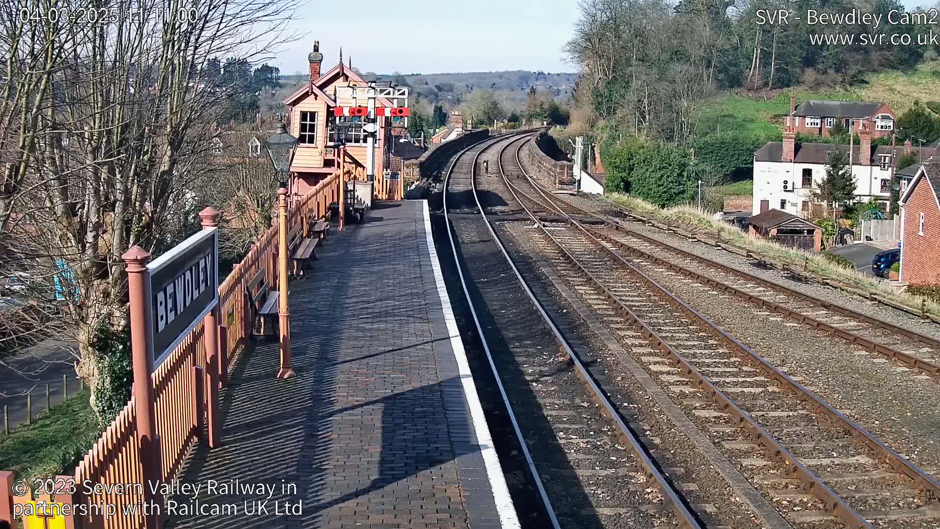 A sunny view of a train station platform with curving train tracks extending into the distance, past houses and trees.