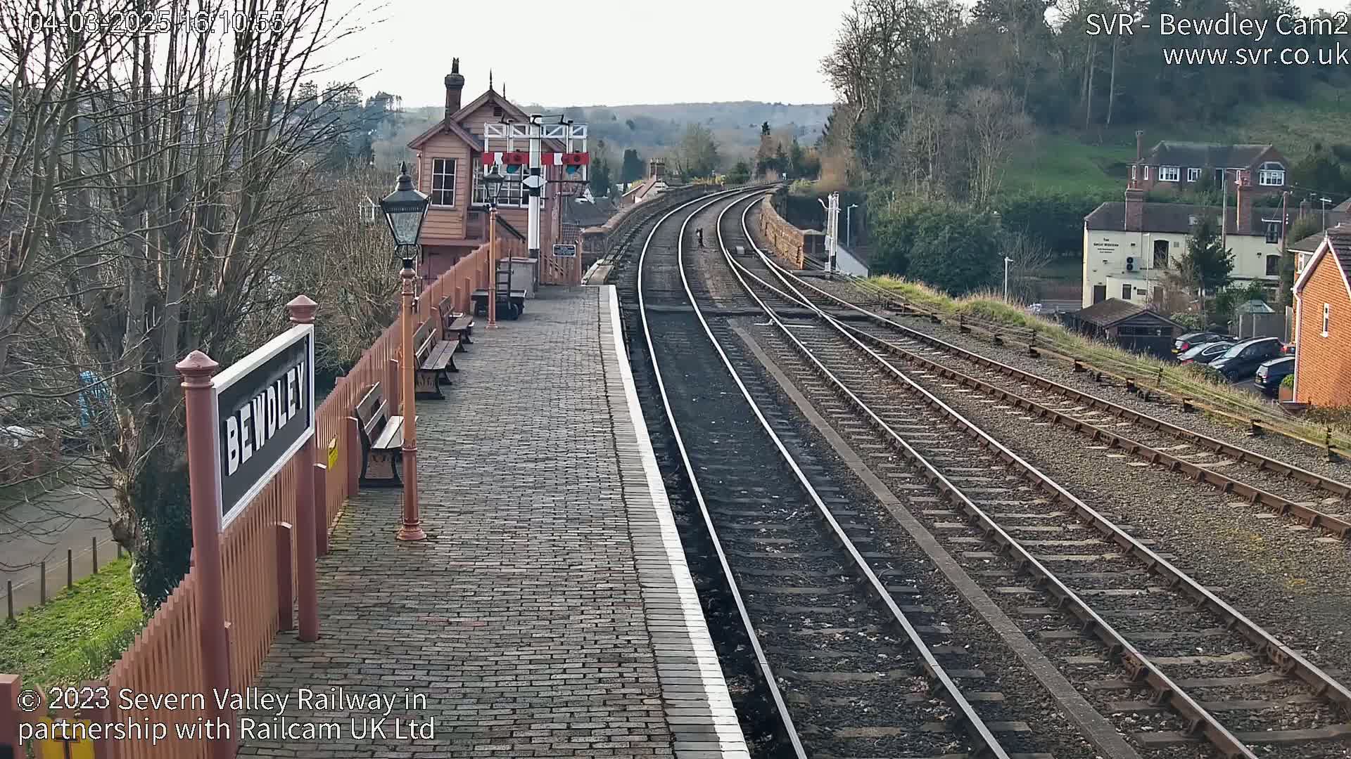 A brick train platform with a signal box and multiple train tracks extends into the distance under a mostly clear sky.