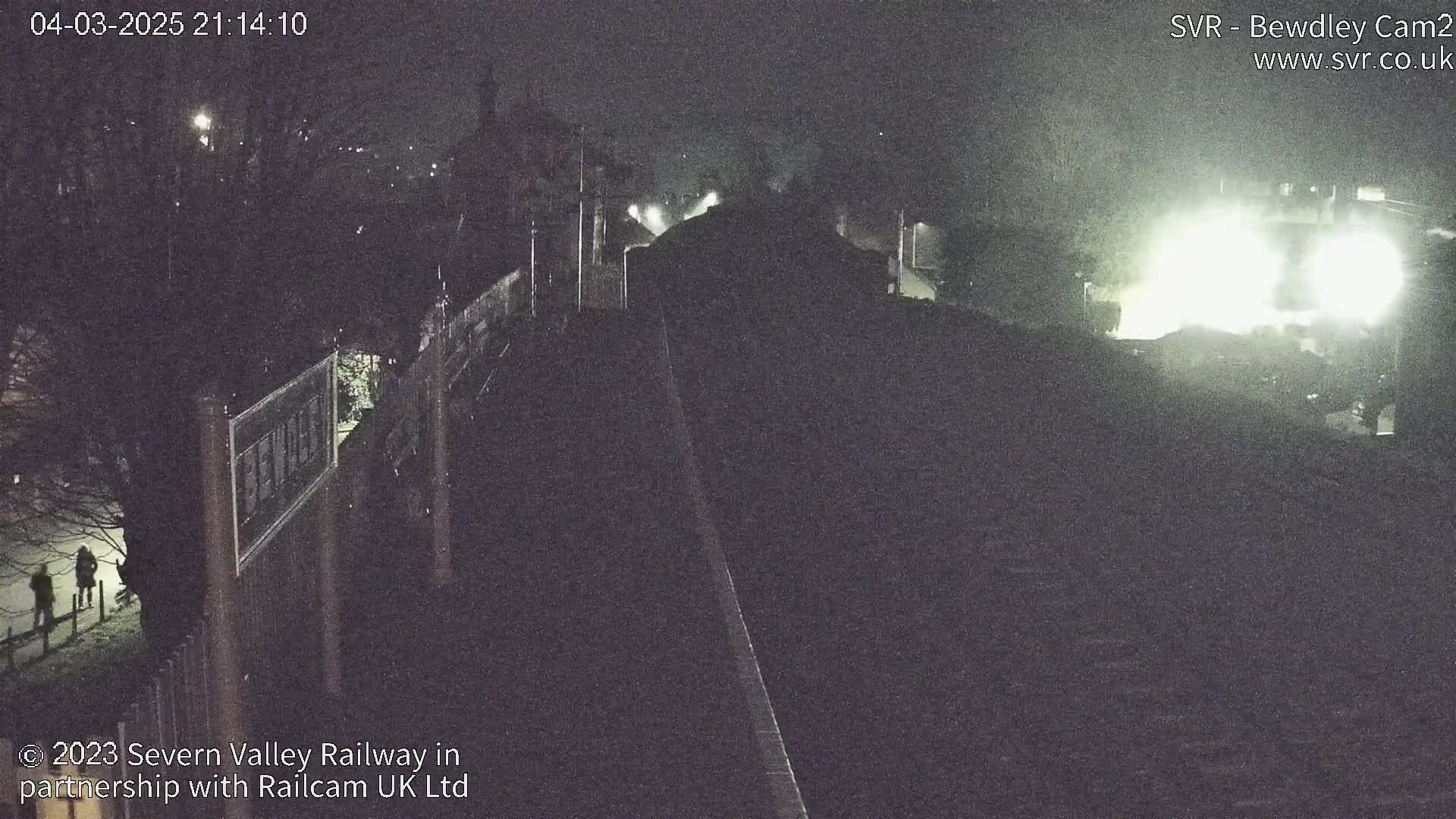 A nighttime, dimly lit view of a railway track and station, with two figures walking on a path to the left.