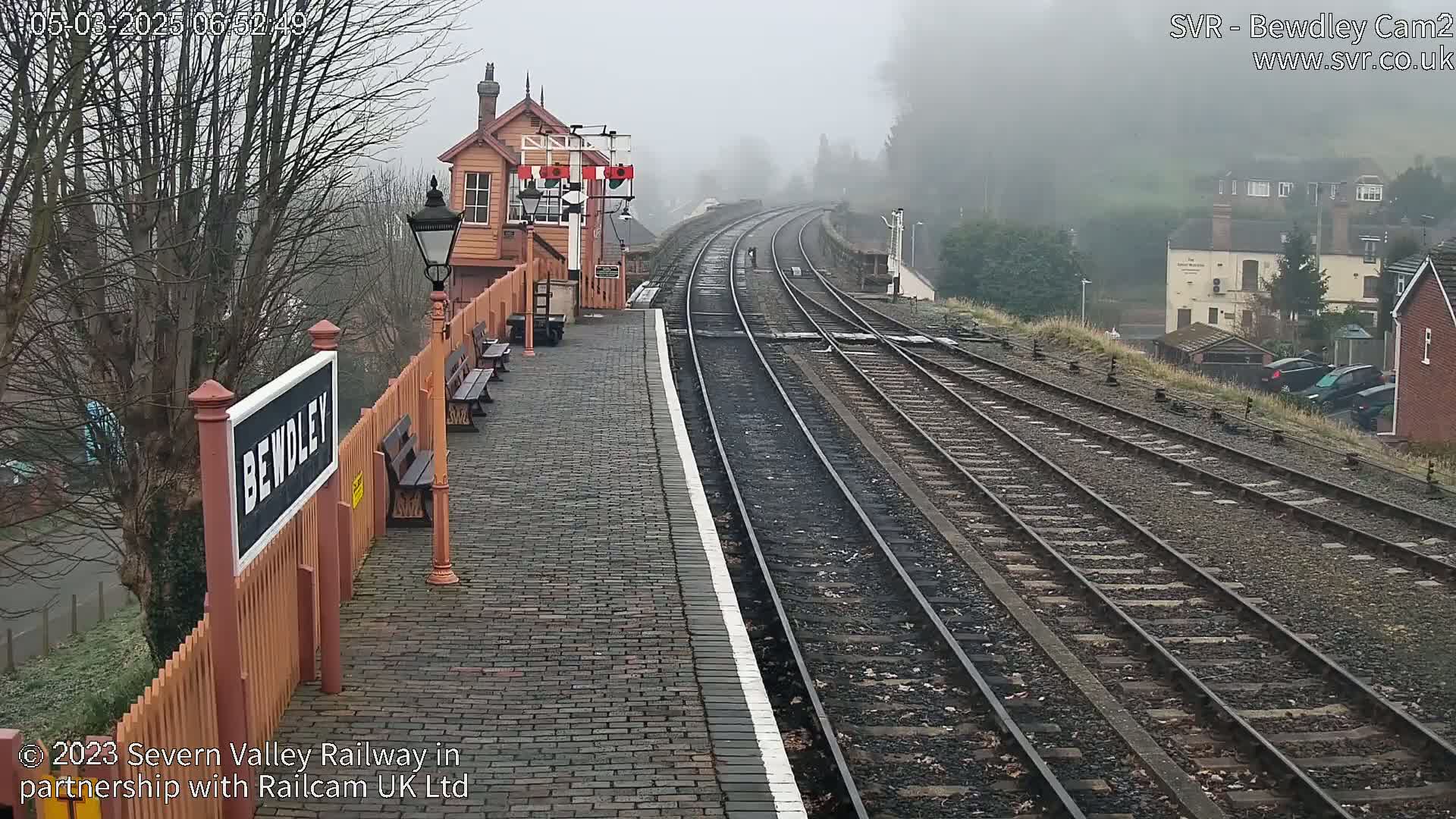 A foggy view of a train station platform with multiple train tracks extending into the distance, a signal box, and several parked cars visible in the background.