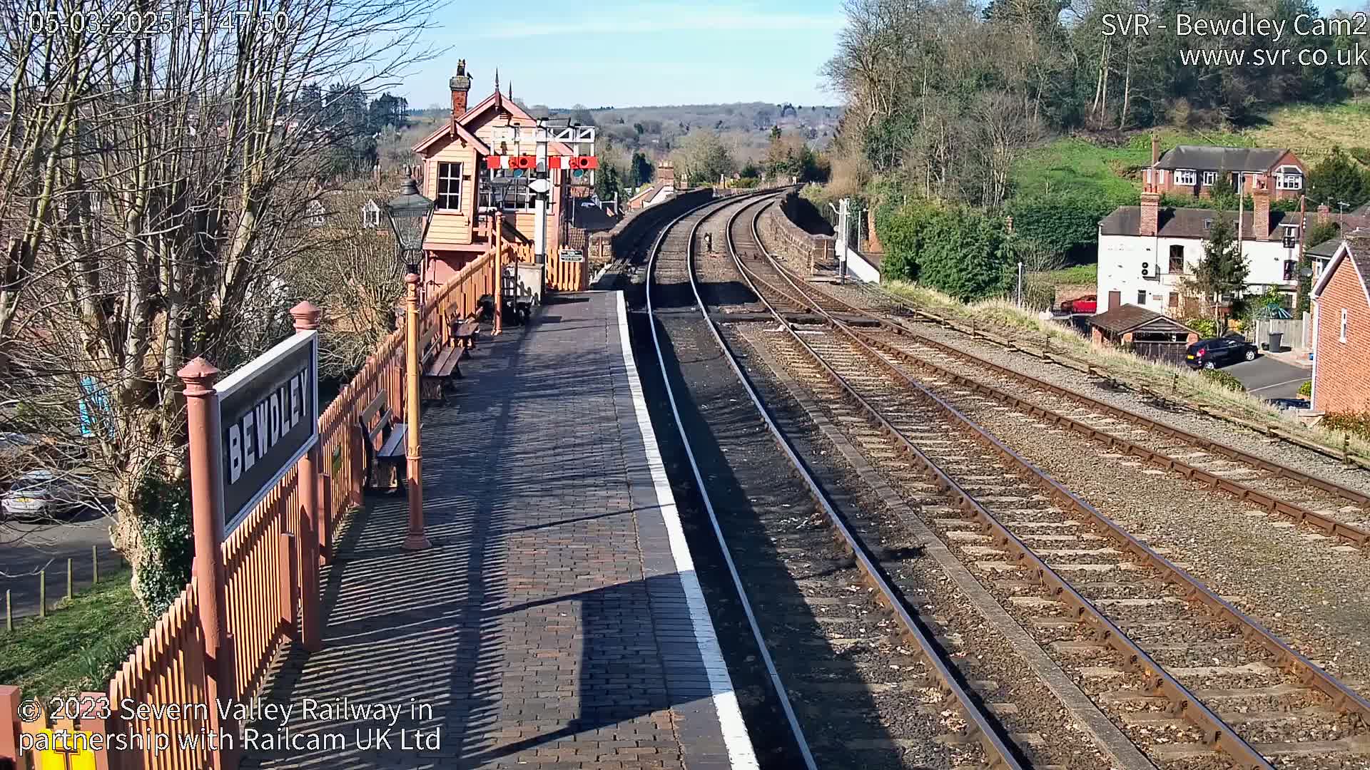 A sunny view of a train station platform with two sets of train tracks curving into the distance, a signal box, and residential buildings beyond.