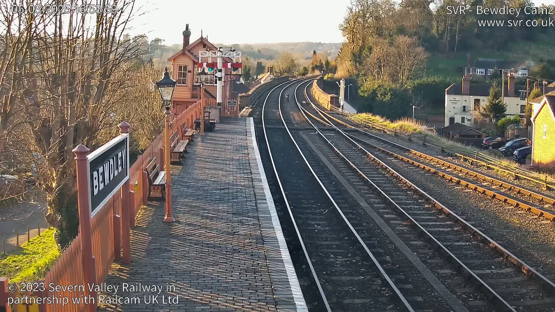A sunny view of a train station platform with multiple train tracks extending into the distance, surrounded by trees and buildings.