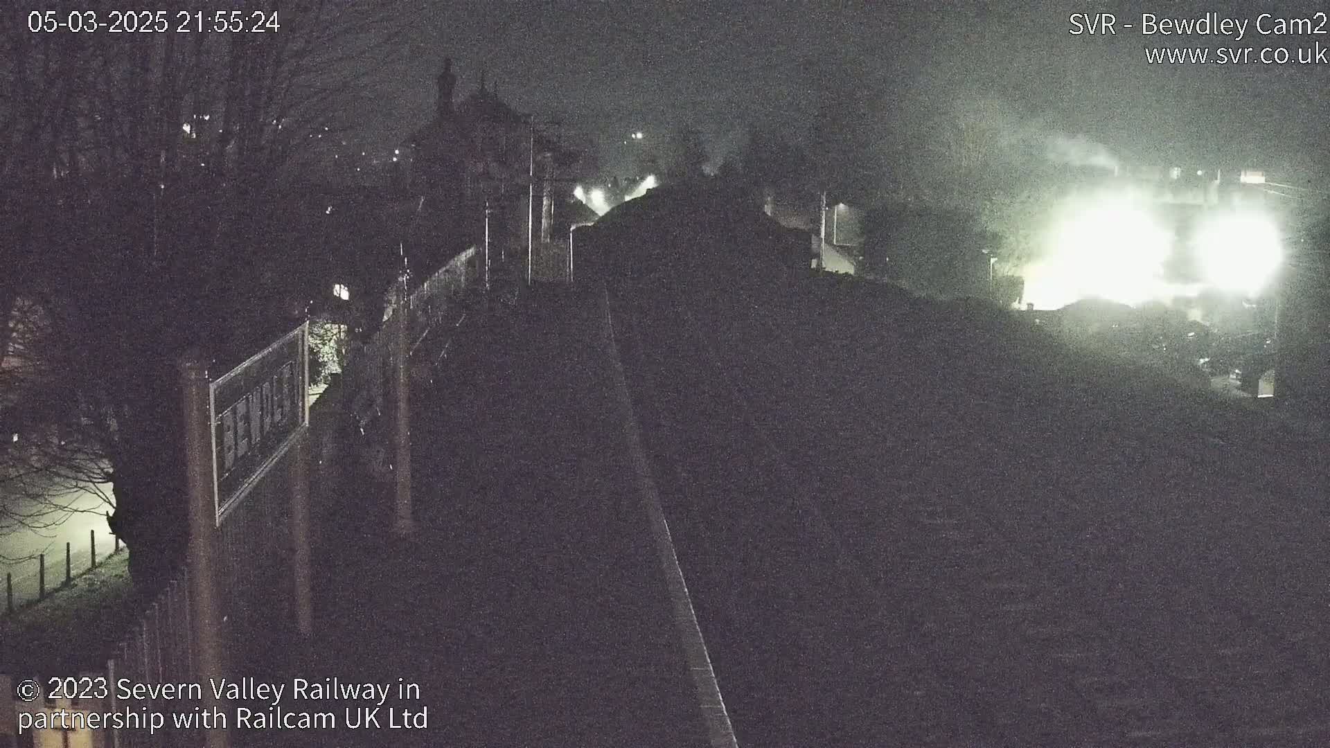 A nighttime view of a dimly lit train station platform and tracks, with foggy or misty conditions.