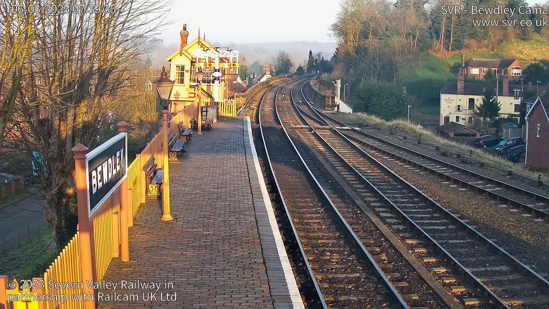 A sunny morning view of a train station platform with several sets of train tracks extending into the distance, past houses and trees on a hillside.