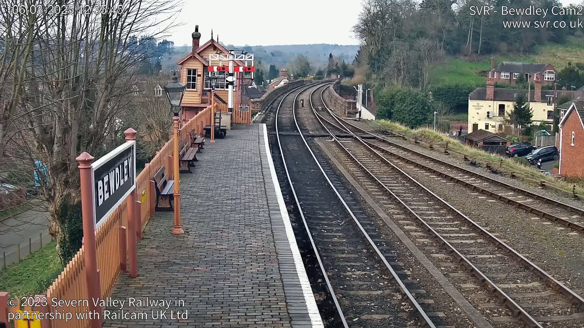 A train station platform with a brick surface and signal box, alongside several train tracks curving into the distance under an overcast sky.