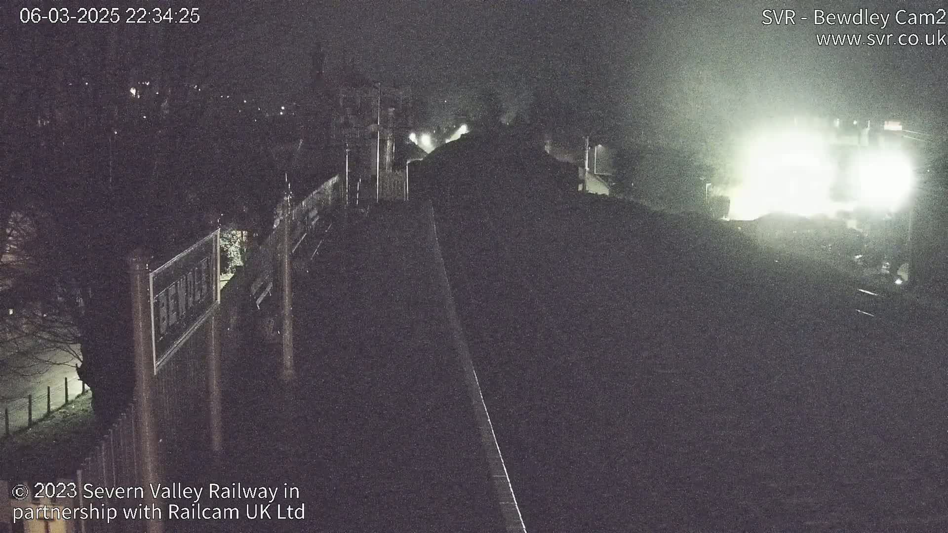 A nighttime, dimly lit view of a train approaching a station, with a bright light source illuminating the scene.