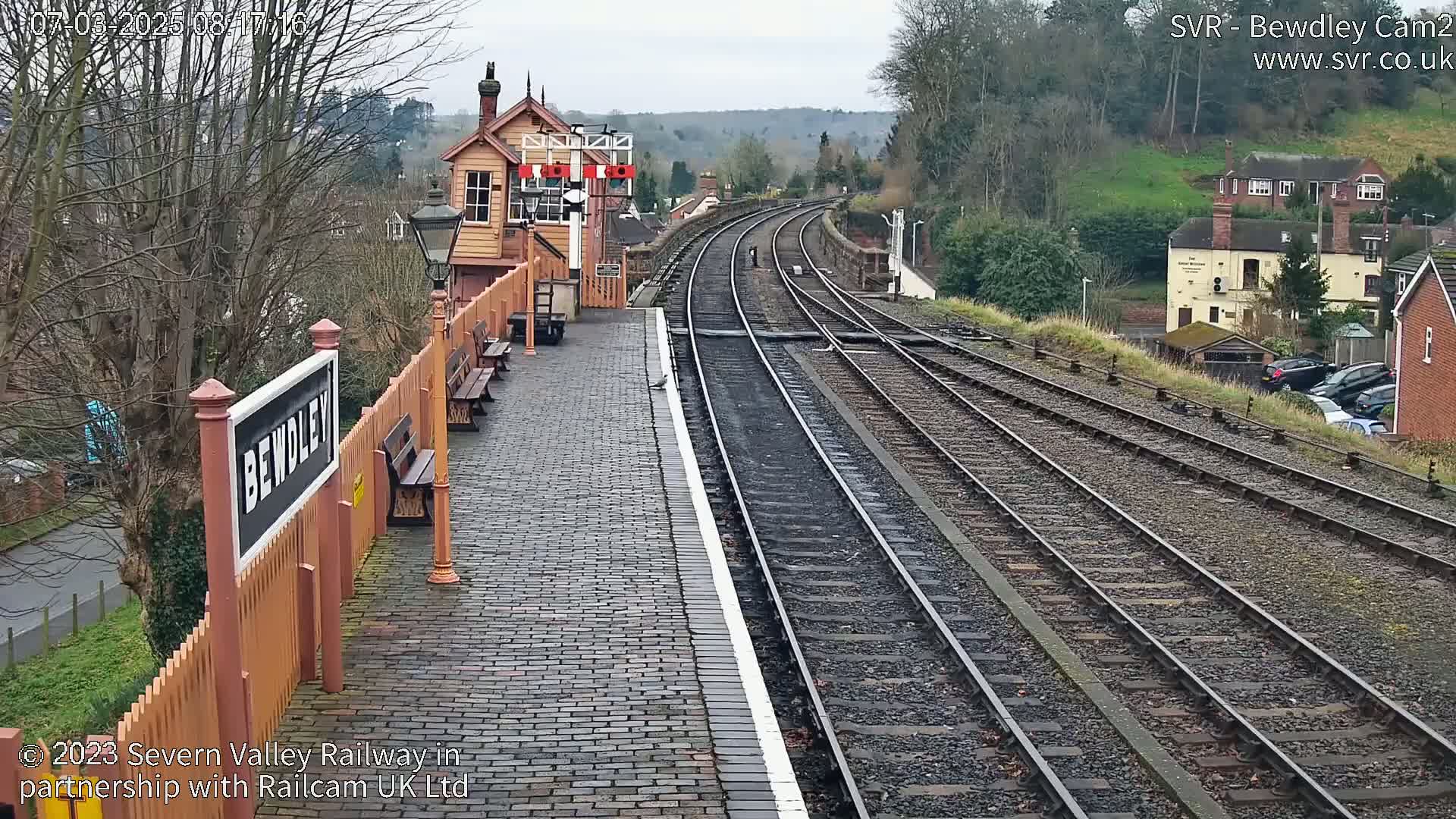A train station platform with a brick surface and several train tracks extending into the distance under an overcast sky.