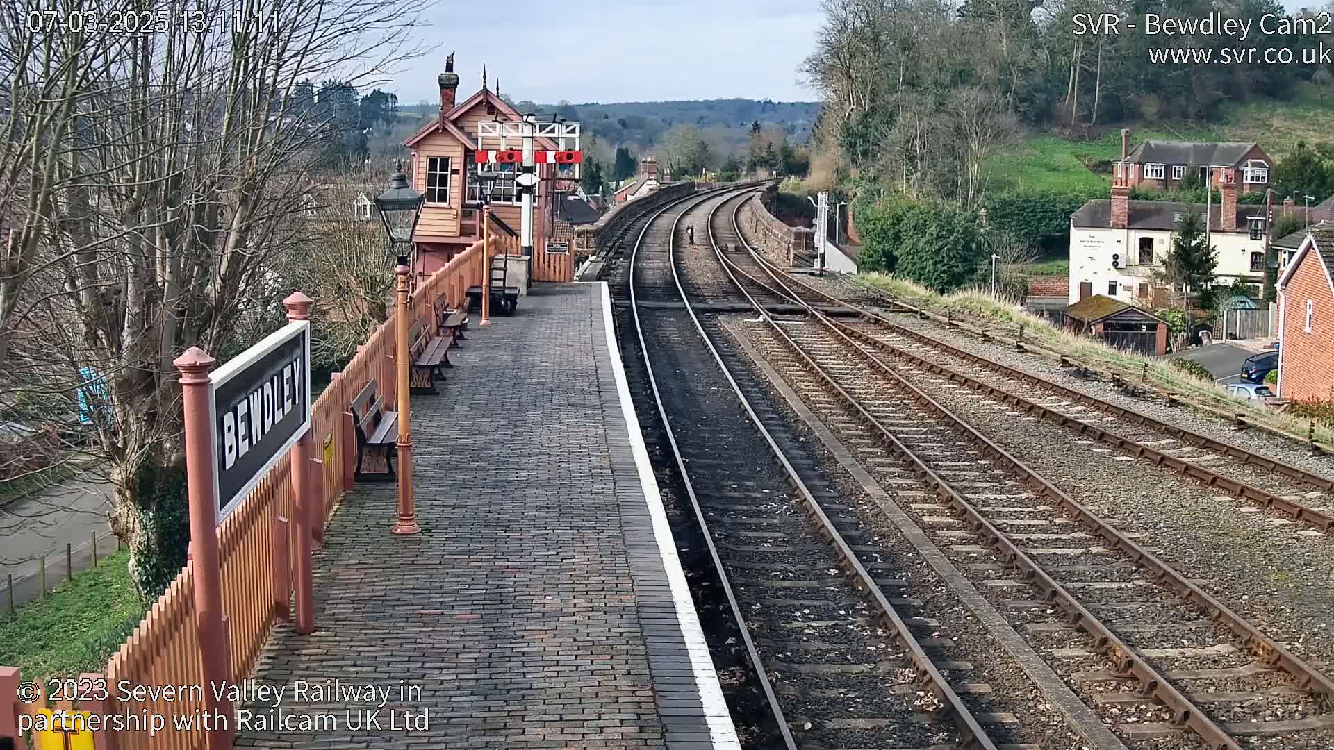 A train station platform with a signal box, and multiple train tracks extending into the distance under a partly sunny sky.