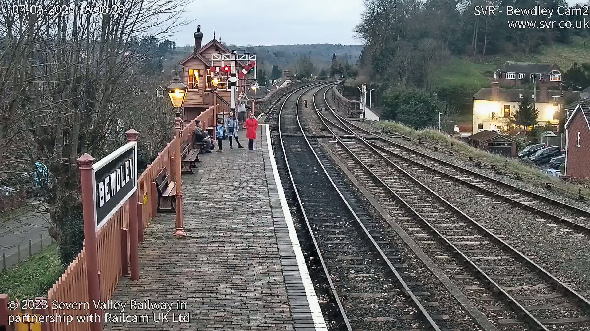 A brick railway platform with several people standing near a sign, overlooking several train tracks that curve into the distance under a cloudy, dusky sky.