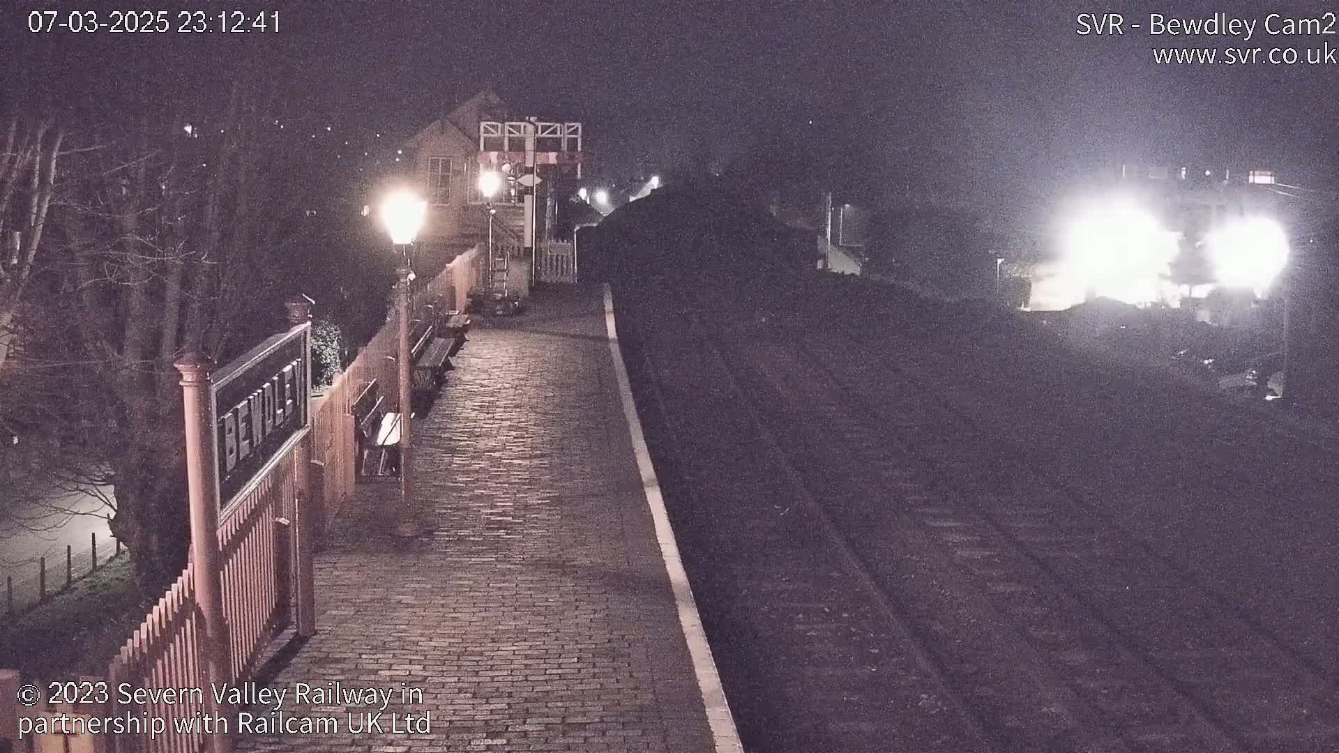 A nighttime view of a train station platform with a train approaching, illuminated by bright headlights under dark skies.