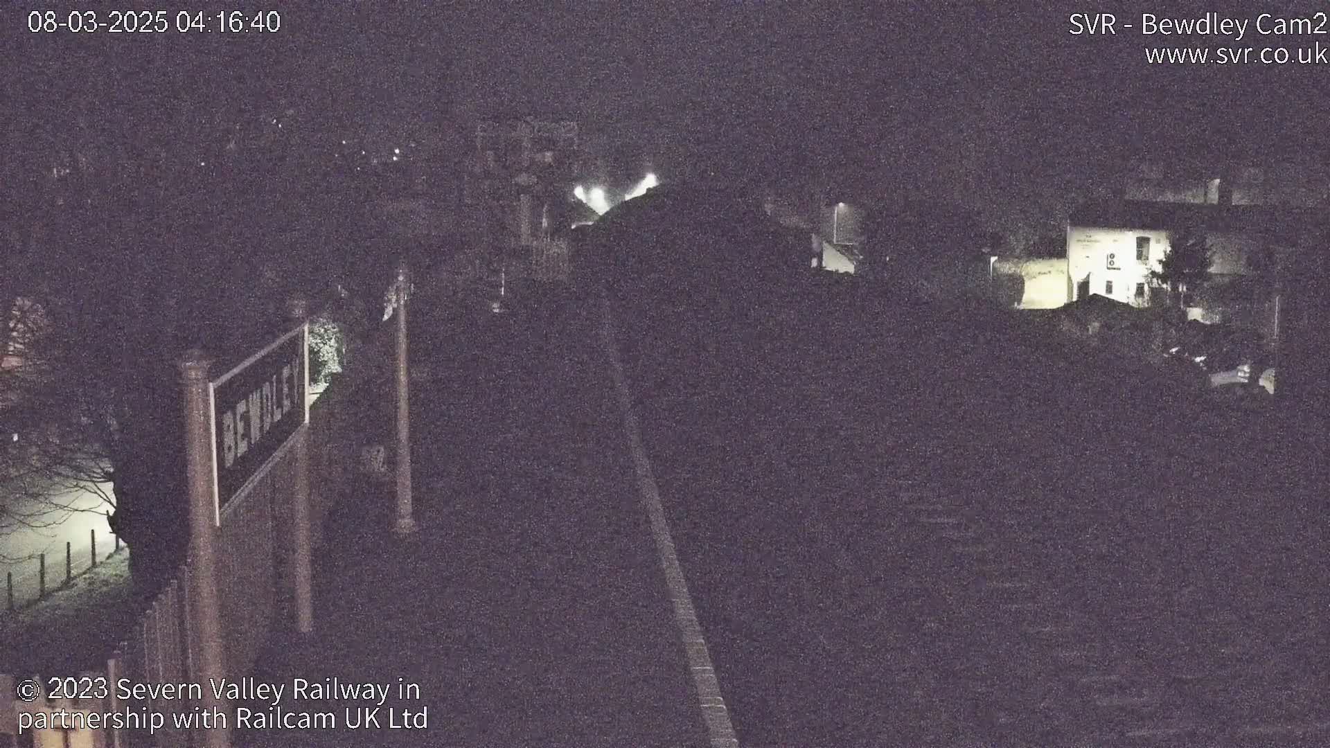 A nighttime, low-light view of a railway line and station, with a partially visible sign and buildings in the background.