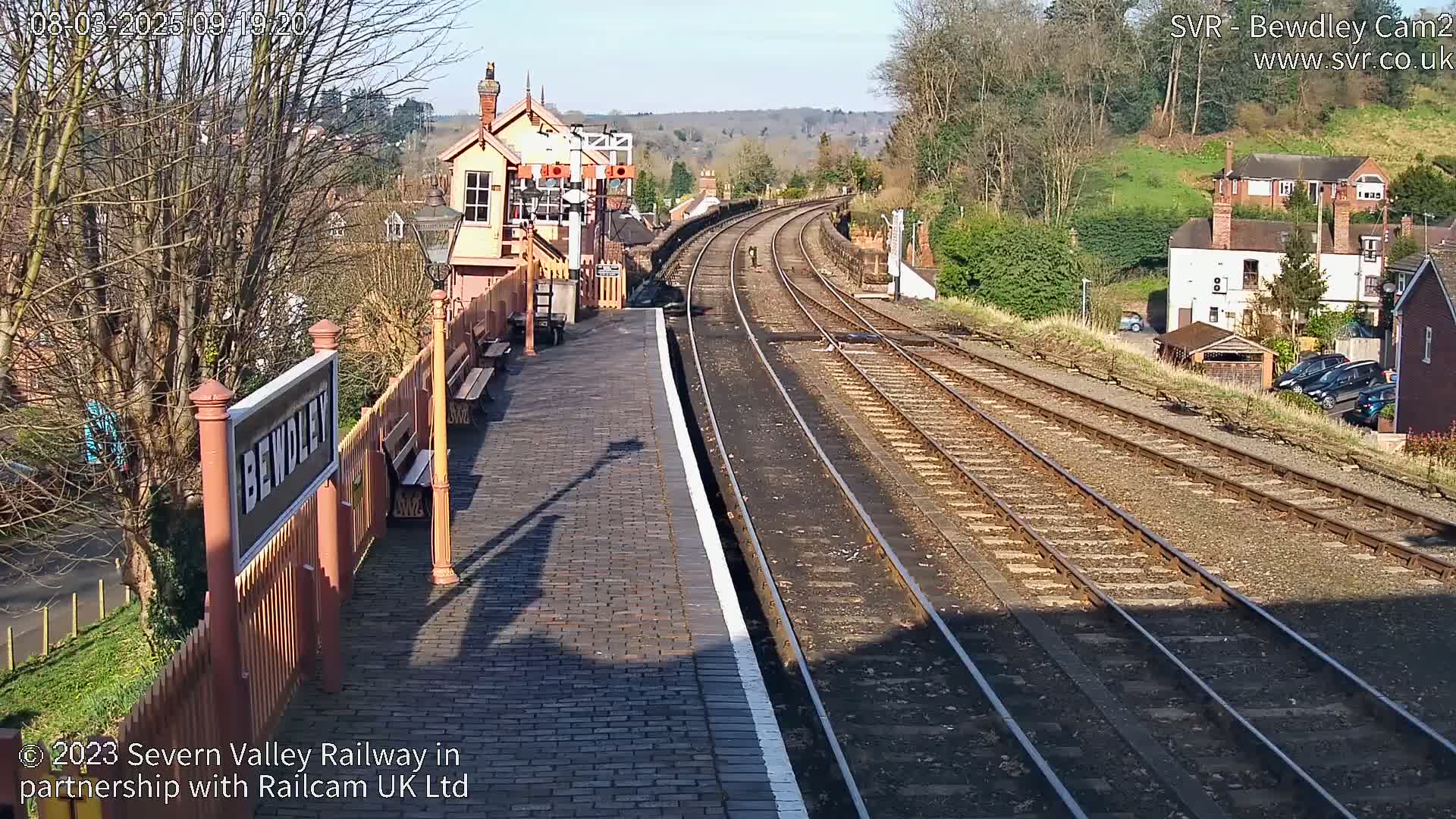 A sunny view of a train station platform with several tracks curving into the distance, surrounded by buildings and trees.